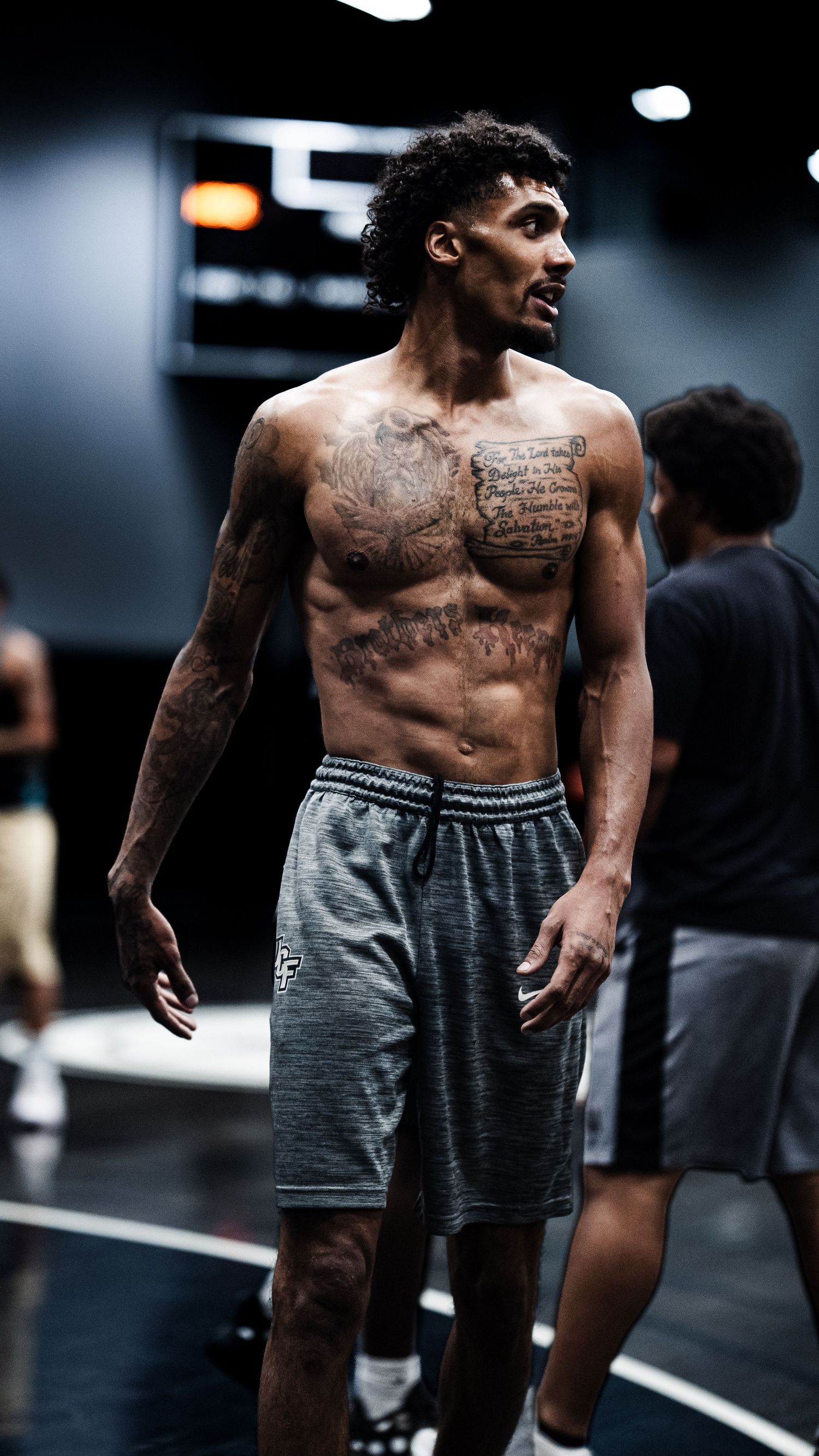 Shirtless athletic man with chest tattoos wearing gray shorts stands confidently on a basketball court.