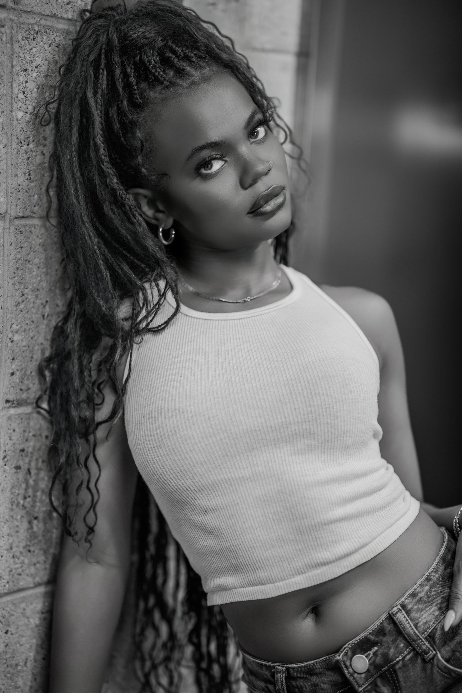 Black and white portrait of a young woman with long braids wearing a white crop top leaning against a wall.