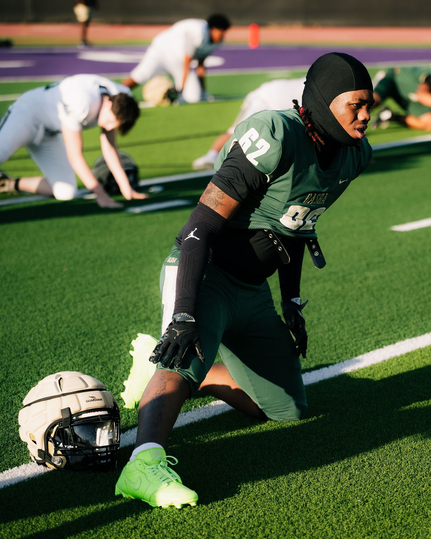 Football player in green uniform stretches on field during pre-game warmups with teammates in background.