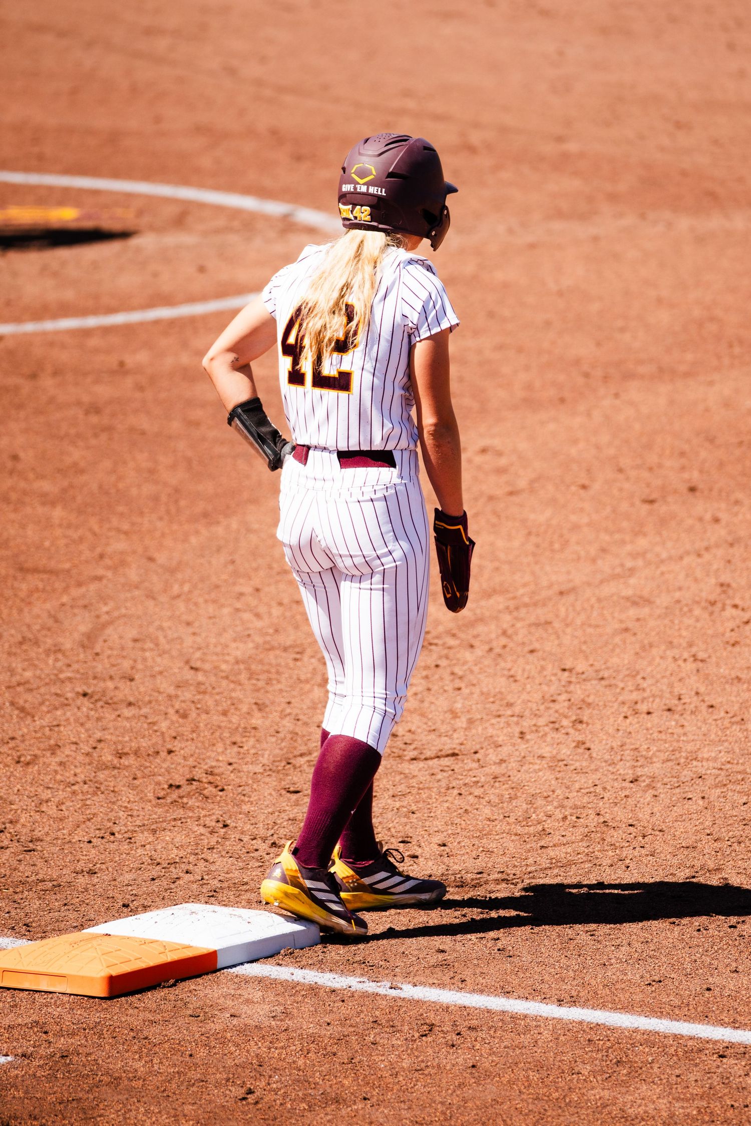 Female softball player #42 in maroon pinstripe uniform standing on base during a game on a red clay field.