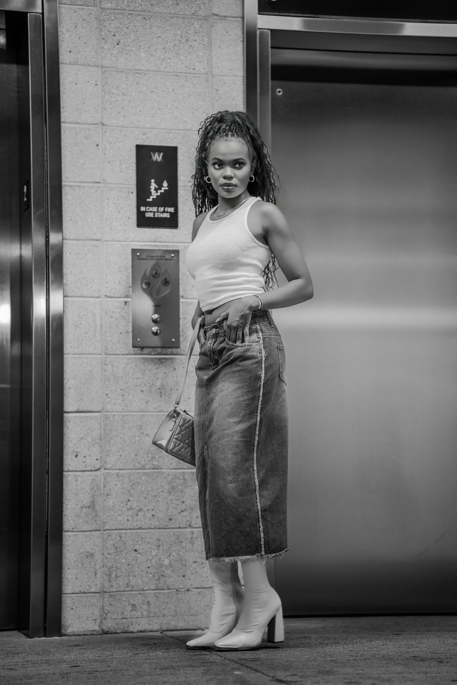 Black and white full-length photo of a fashionable woman in white crop top, denim skirt, and heeled boots by elevators.