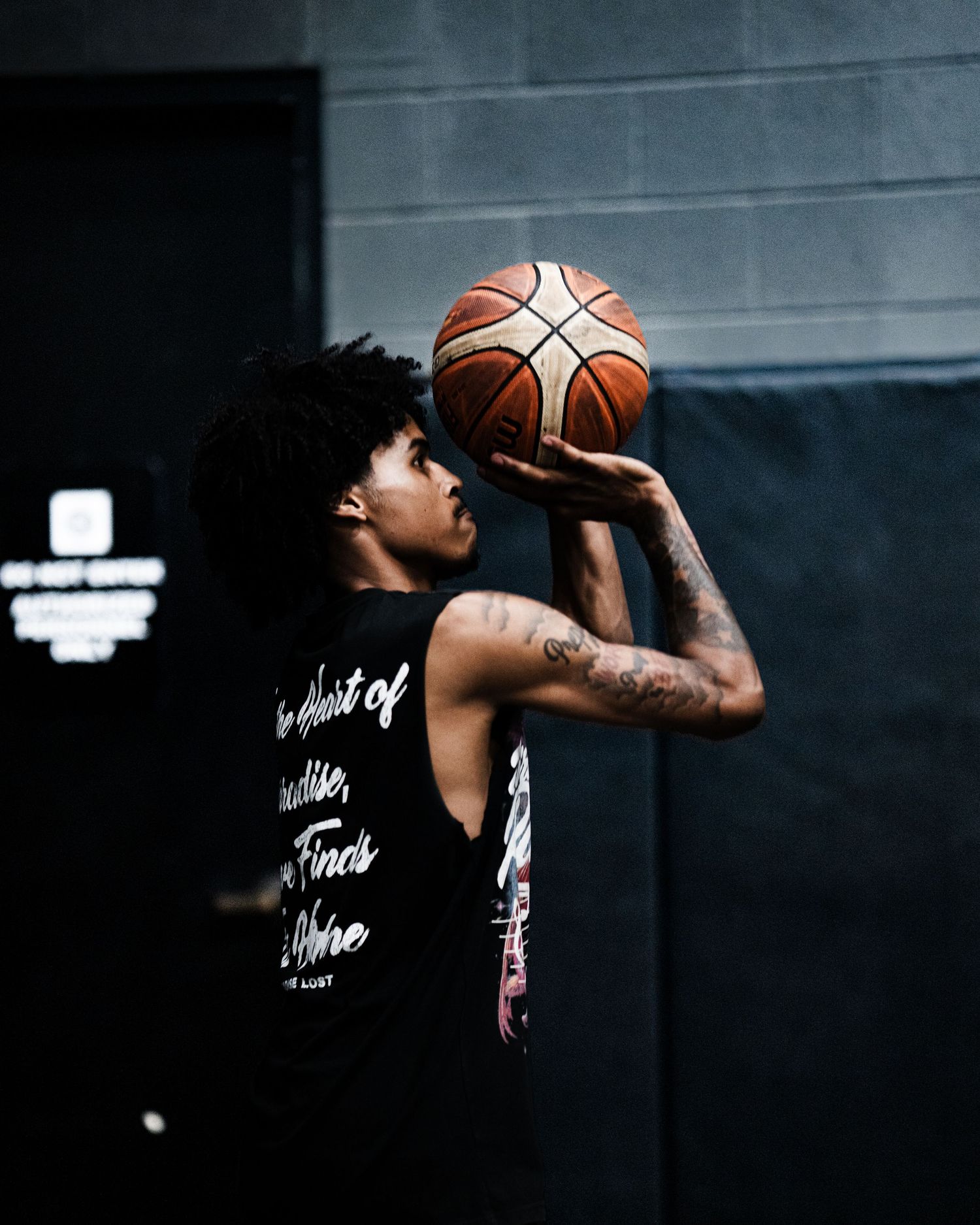 Tattooed basketball player in black sleeveless shirt shoots a ball indoors at a gym.