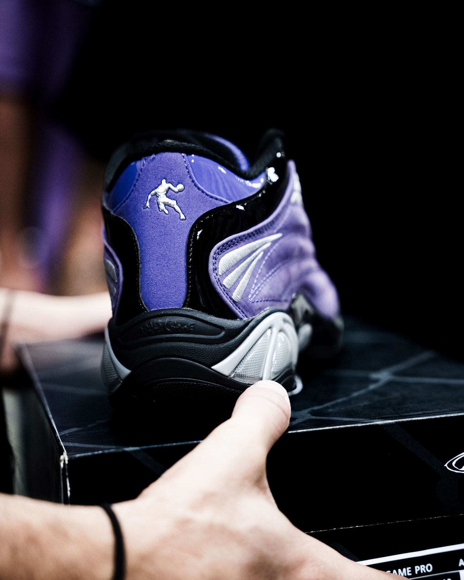 Purple and black Nike basketball sneaker displayed on its box, held by a hand against a dark background.