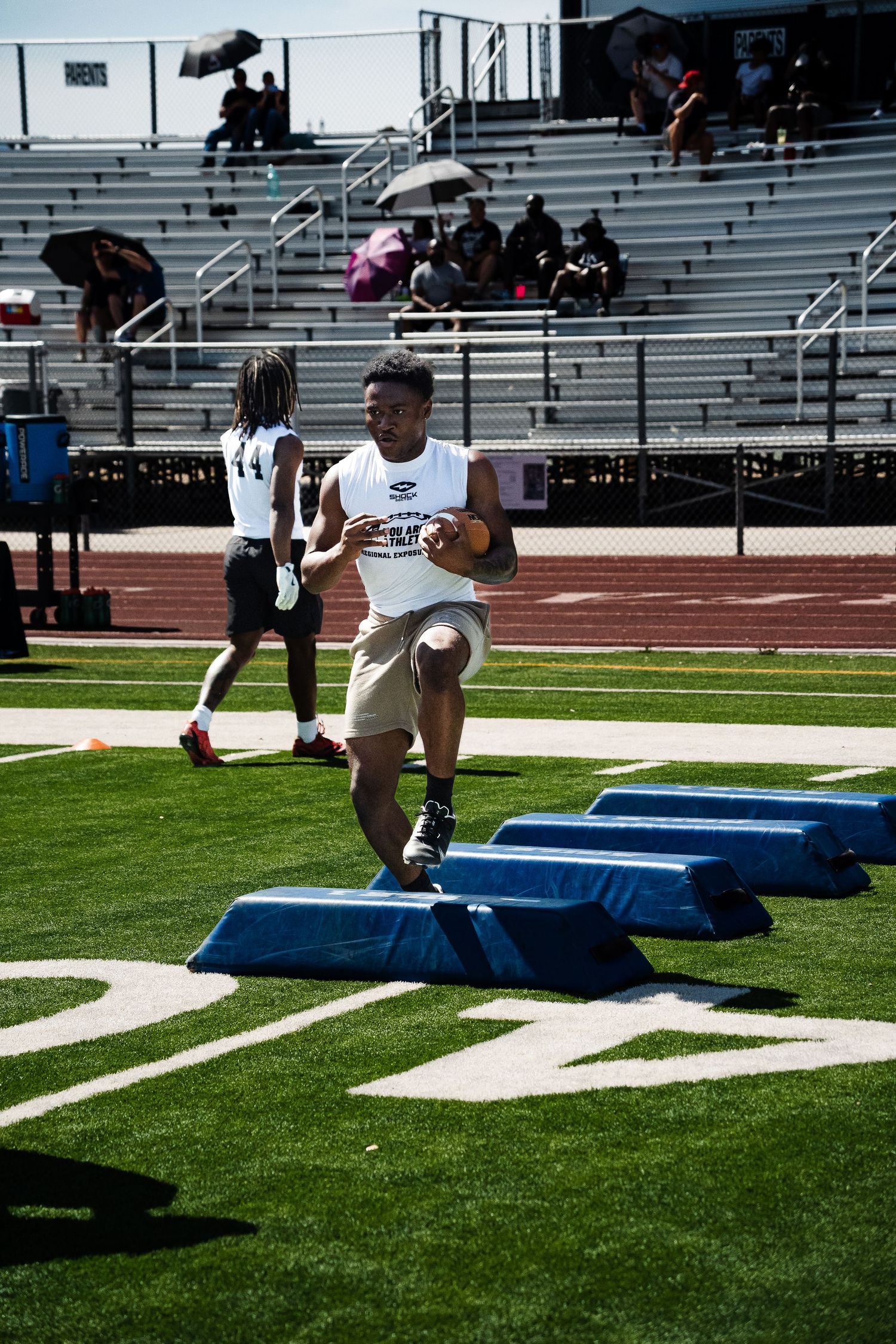 Young football player runs agility drill over blue barriers on sunny stadium field.