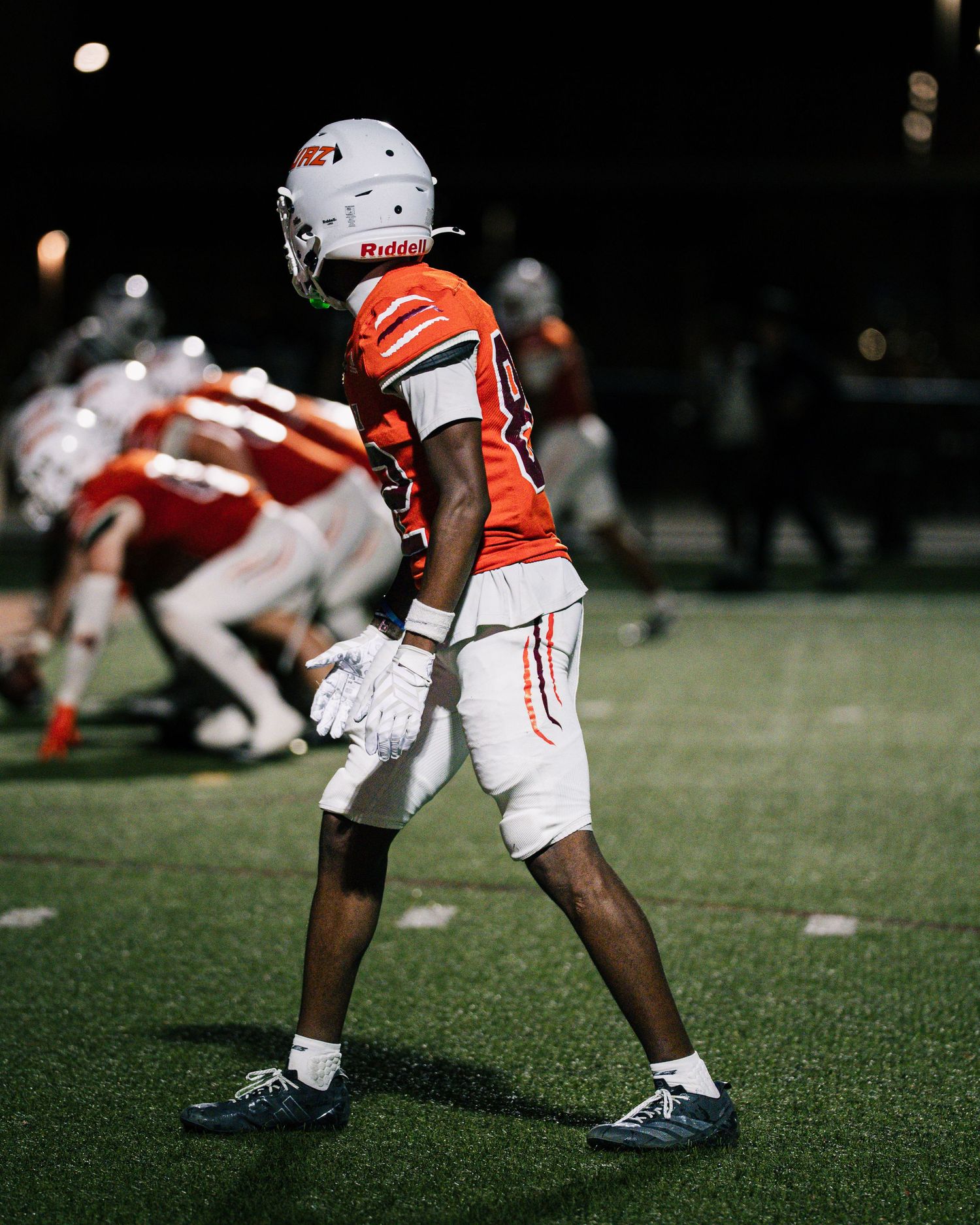 A football player in an orange uniform and white helmet stands on a lit field during a night game.