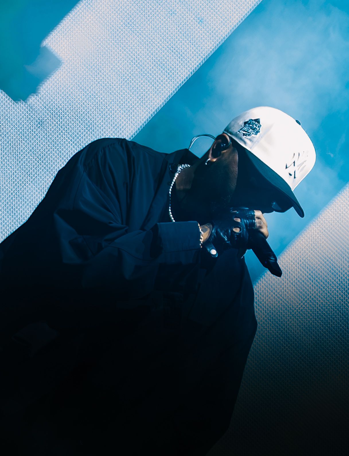 Performer in a white cap, dark oversized jacket and black gloves leans forward on stage, bathed in blue light and haze.