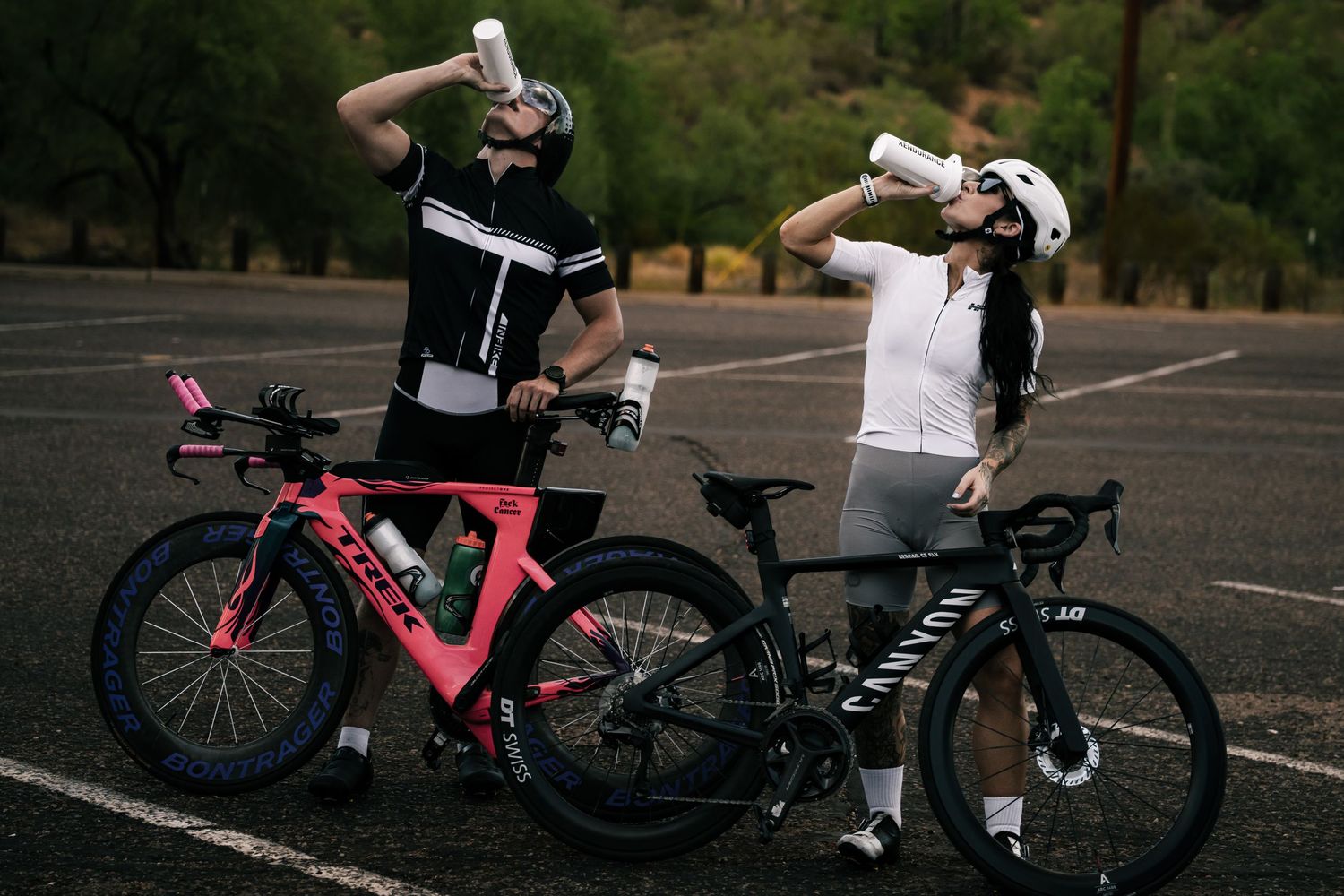 Two cyclists in athletic gear drink from water bottles beside a pink and black Canyon bike in a parking lot.
