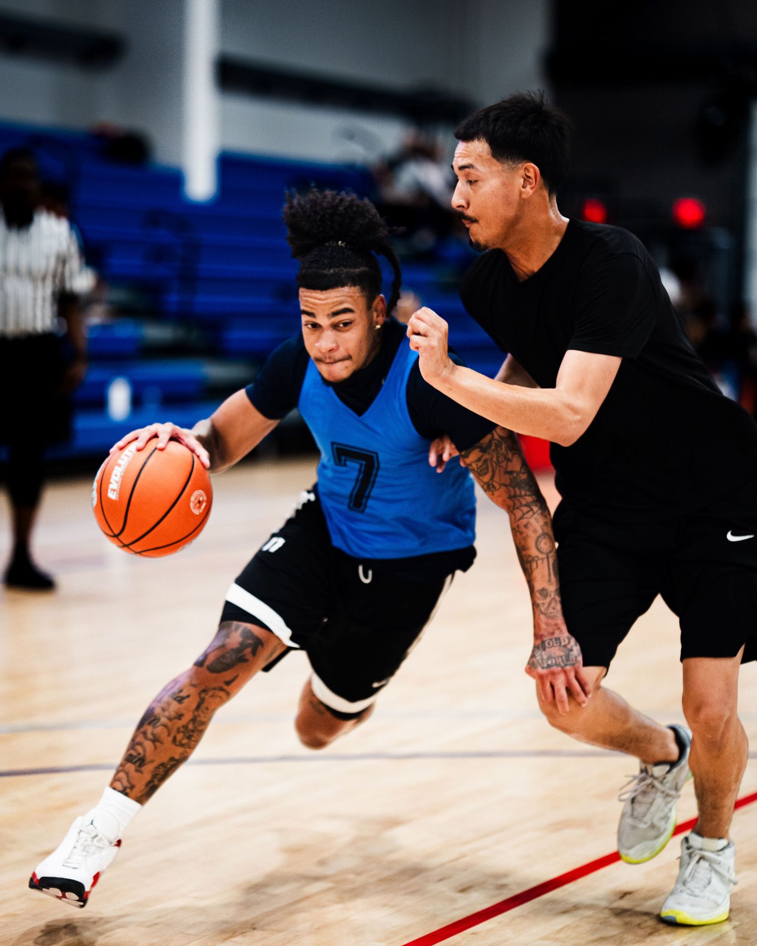 Player #7 in blue jersey dribbles past defender during indoor basketball game on hardwood court.
