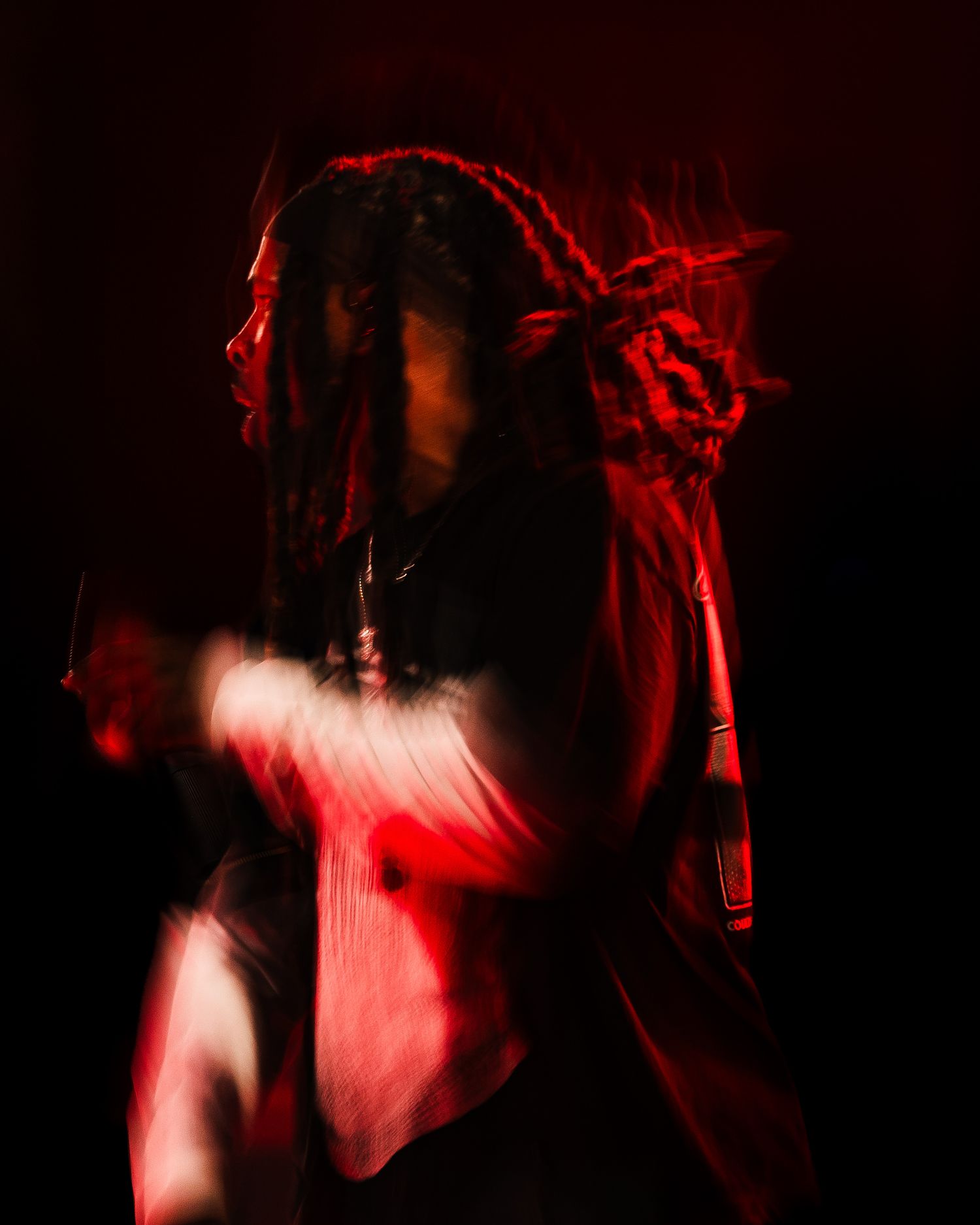 A person with dreadlocks poses dramatically under red lighting creating a blurred motion effect against a dark background.