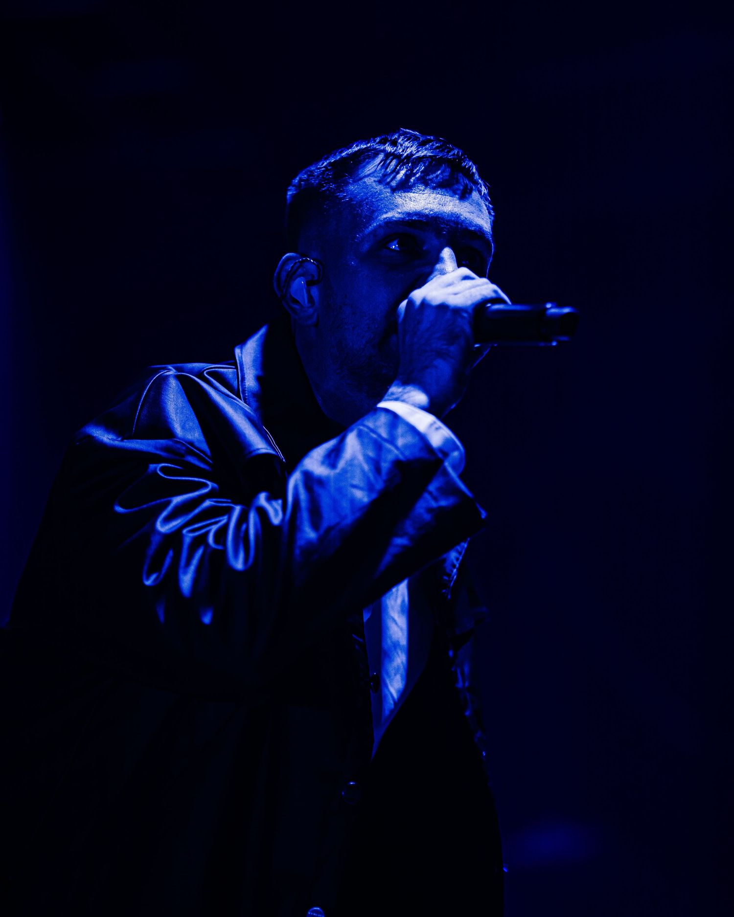 Male performer singing into microphone on stage bathed in dramatic blue lighting wearing a leather jacket.