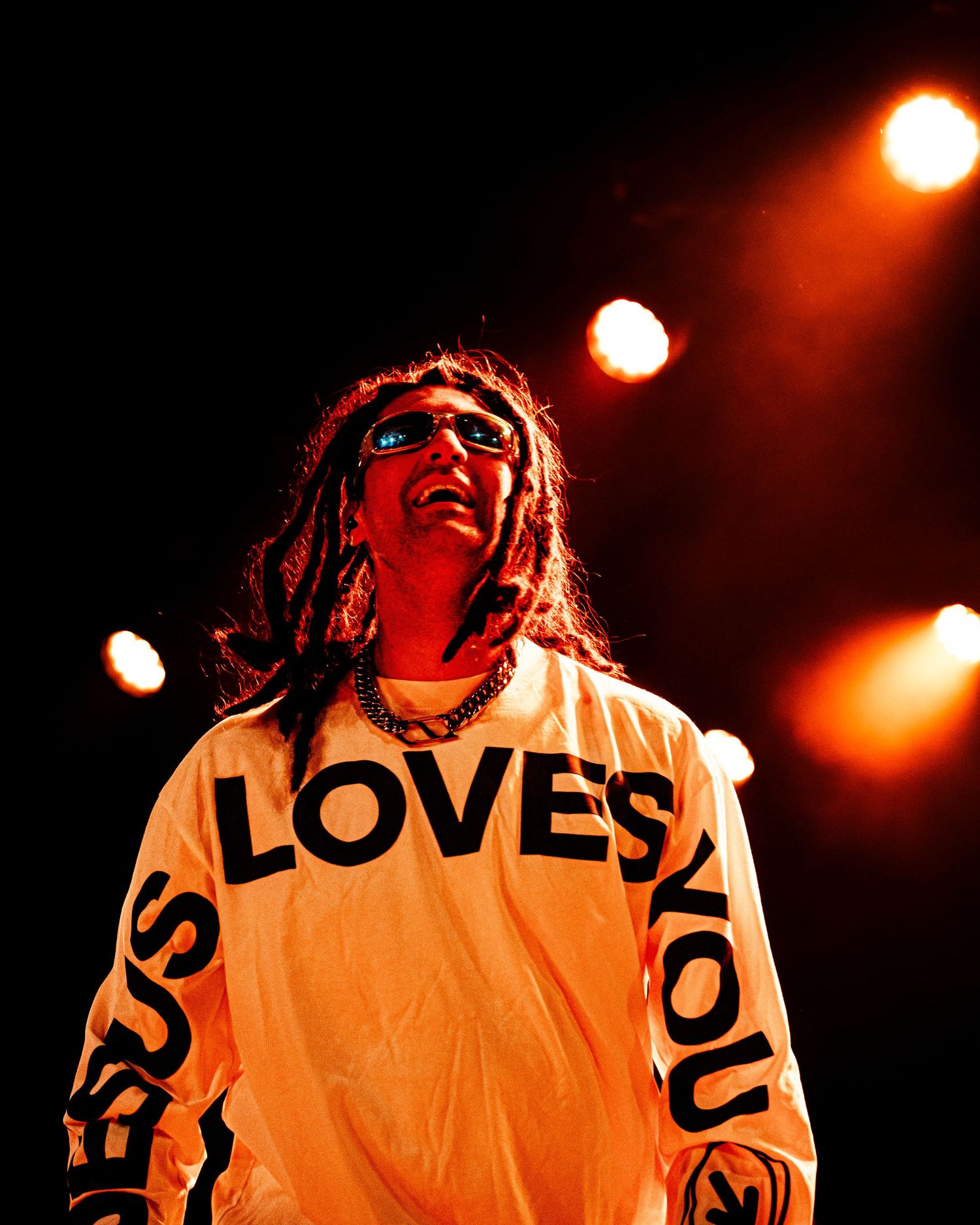 Performer in Jesus Loves You shirt on stage with dramatic orange lighting and long dark hair.