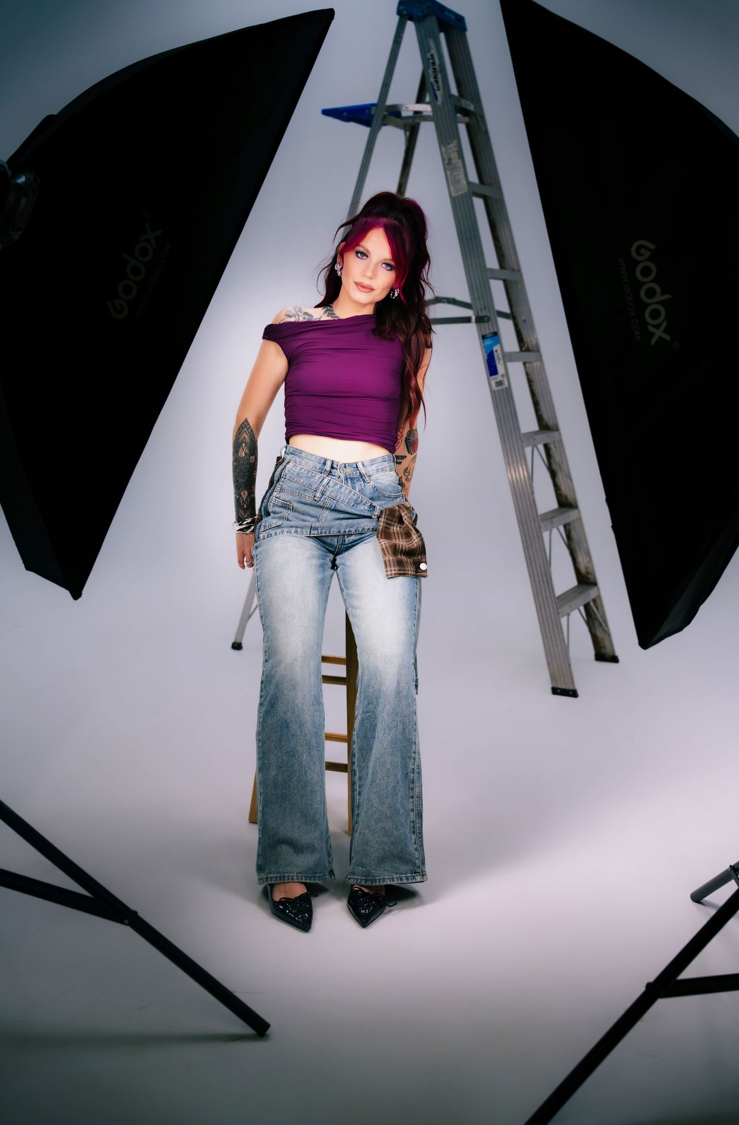 Tattooed model with purple hair wearing a purple crop top and flared jeans stands between Godox studio lights.