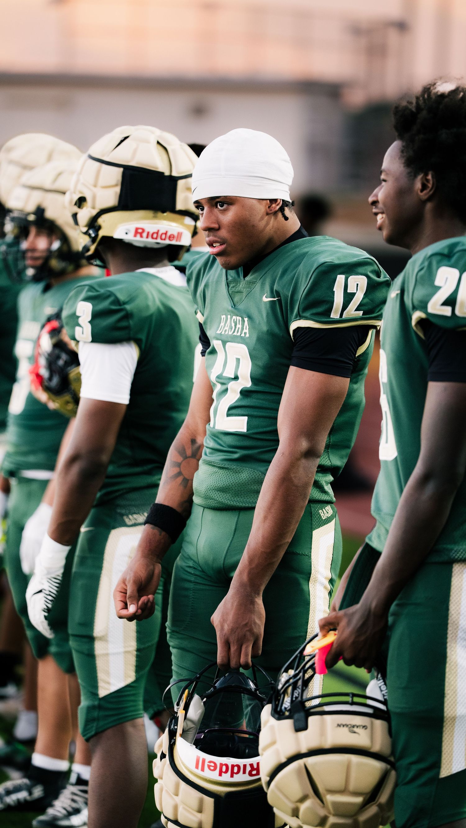 High school football players in green Riddell uniforms stand on the sideline during a game, with player #22 holding his helmet.