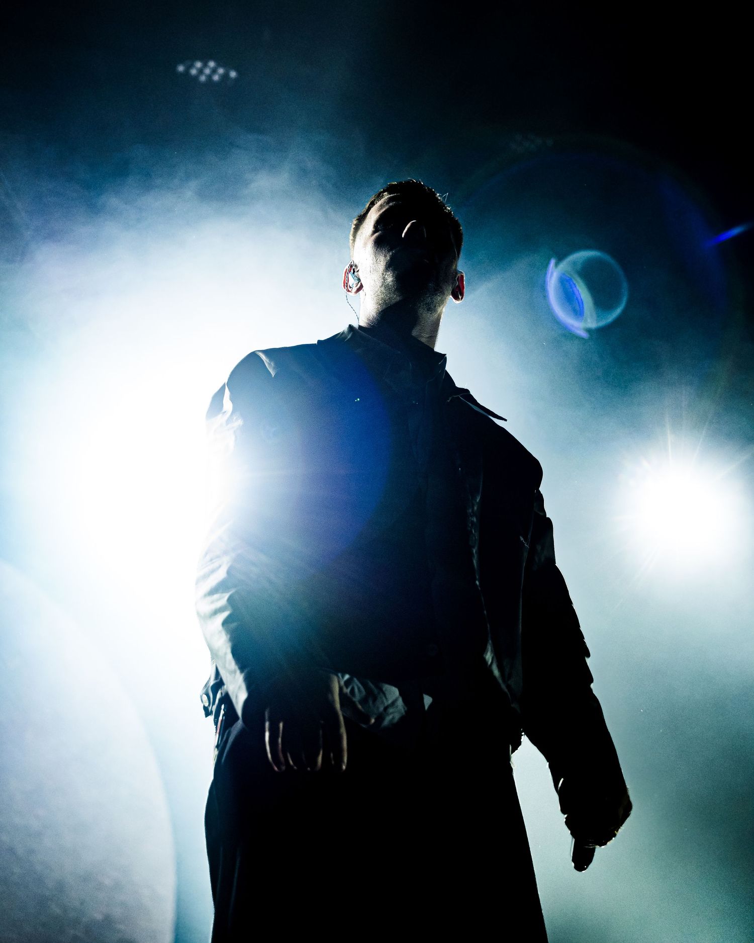 Silhouetted figure in black coat stands dramatically backlit by bright stage lights with smoke and lens flare effects.