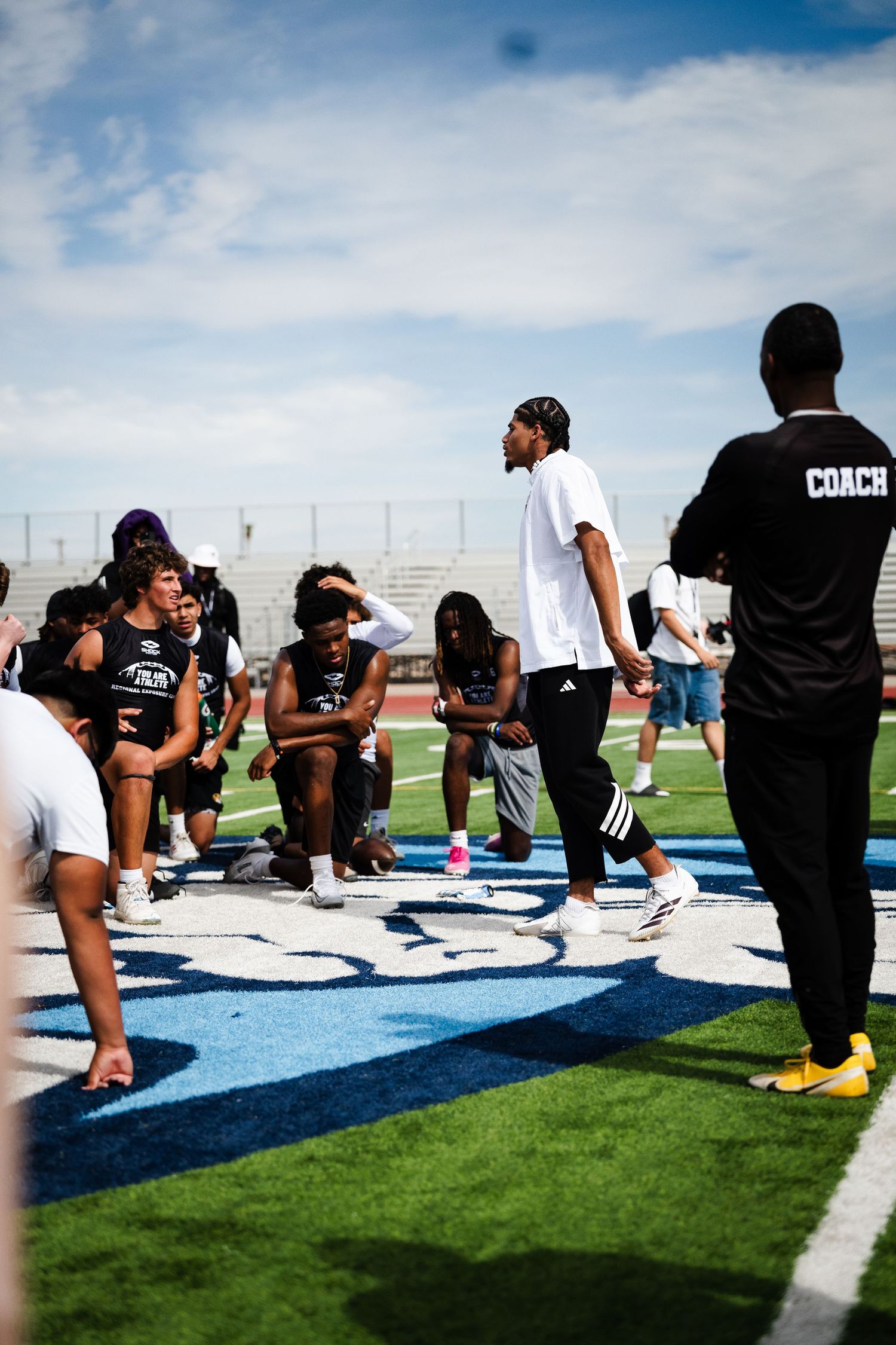 Coach addresses football players sitting on field during outdoor team practice session on blue turf.