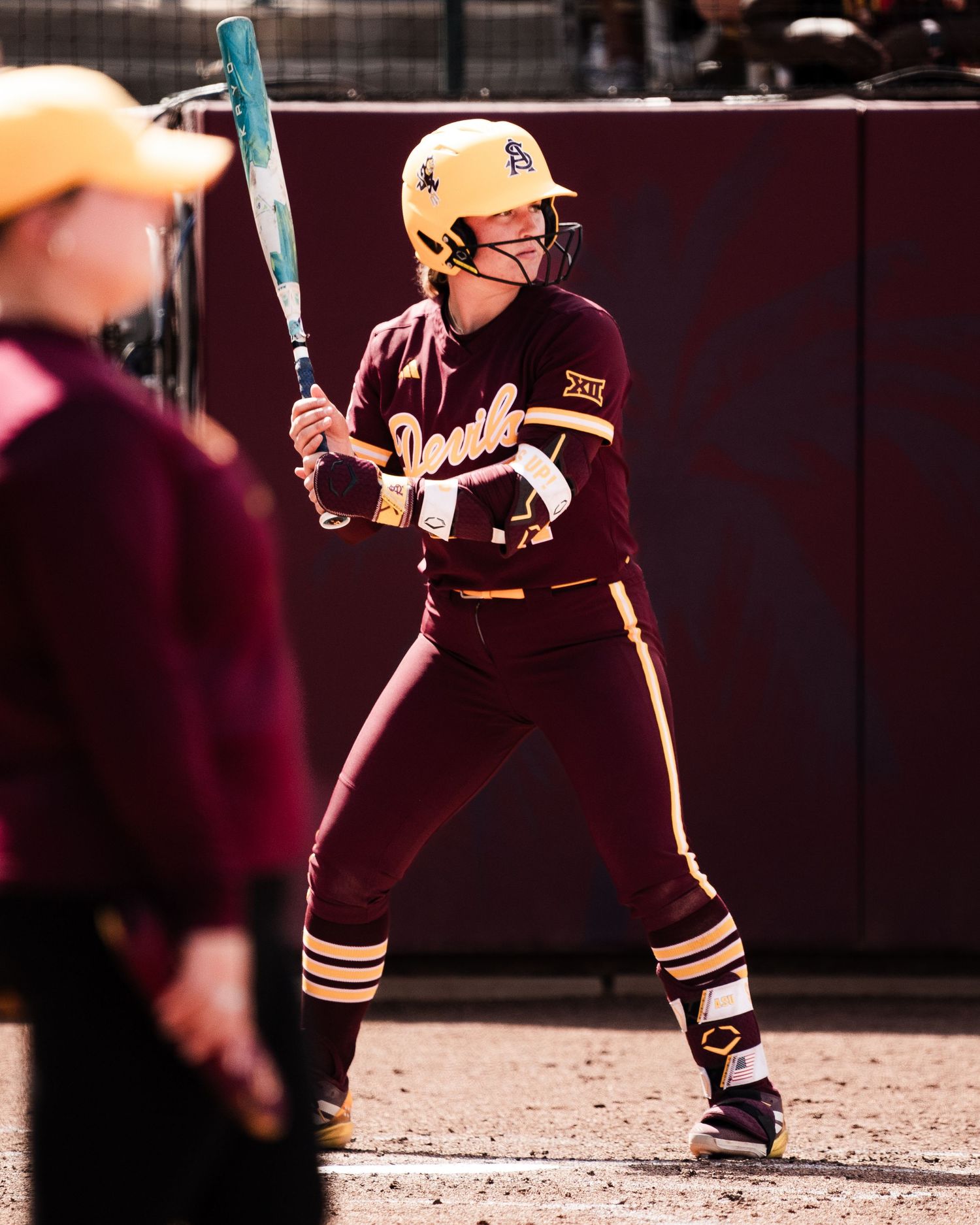 ASU softball player in maroon and gold uniform stands at bat holding a teal bat during a college game.