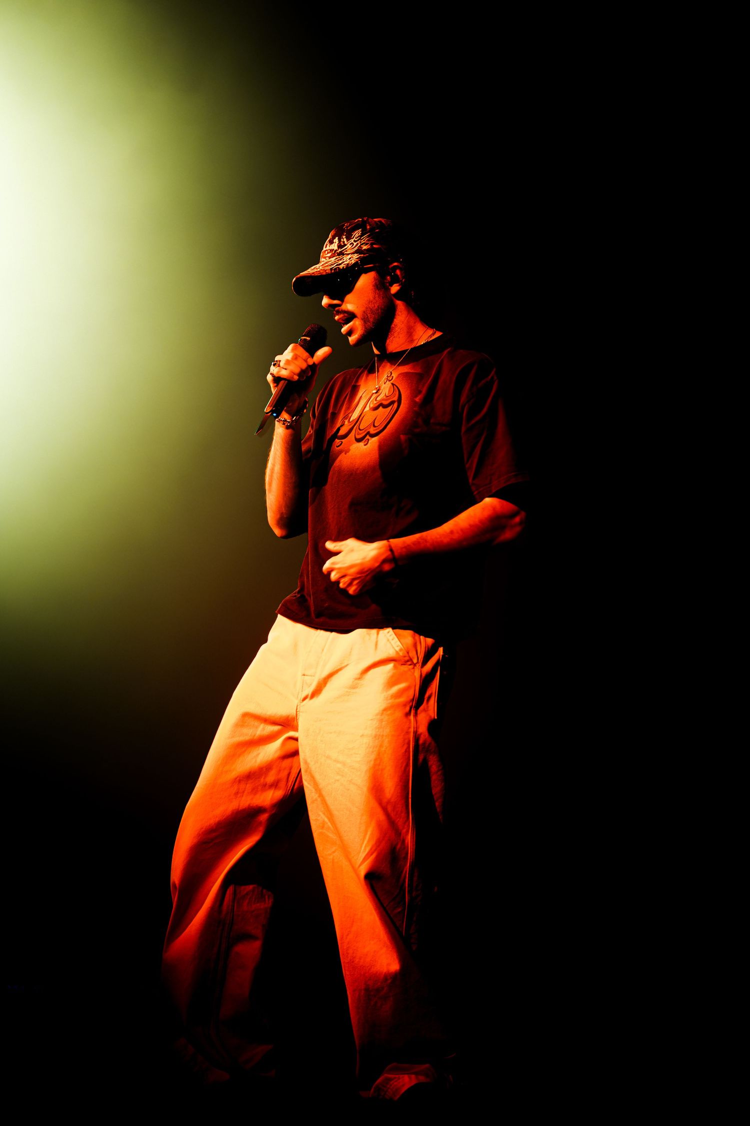 Performer in a patterned cap, sunglasses, maroon t-shirt and light pants sings into a microphone on a dark stage under warm orange light.