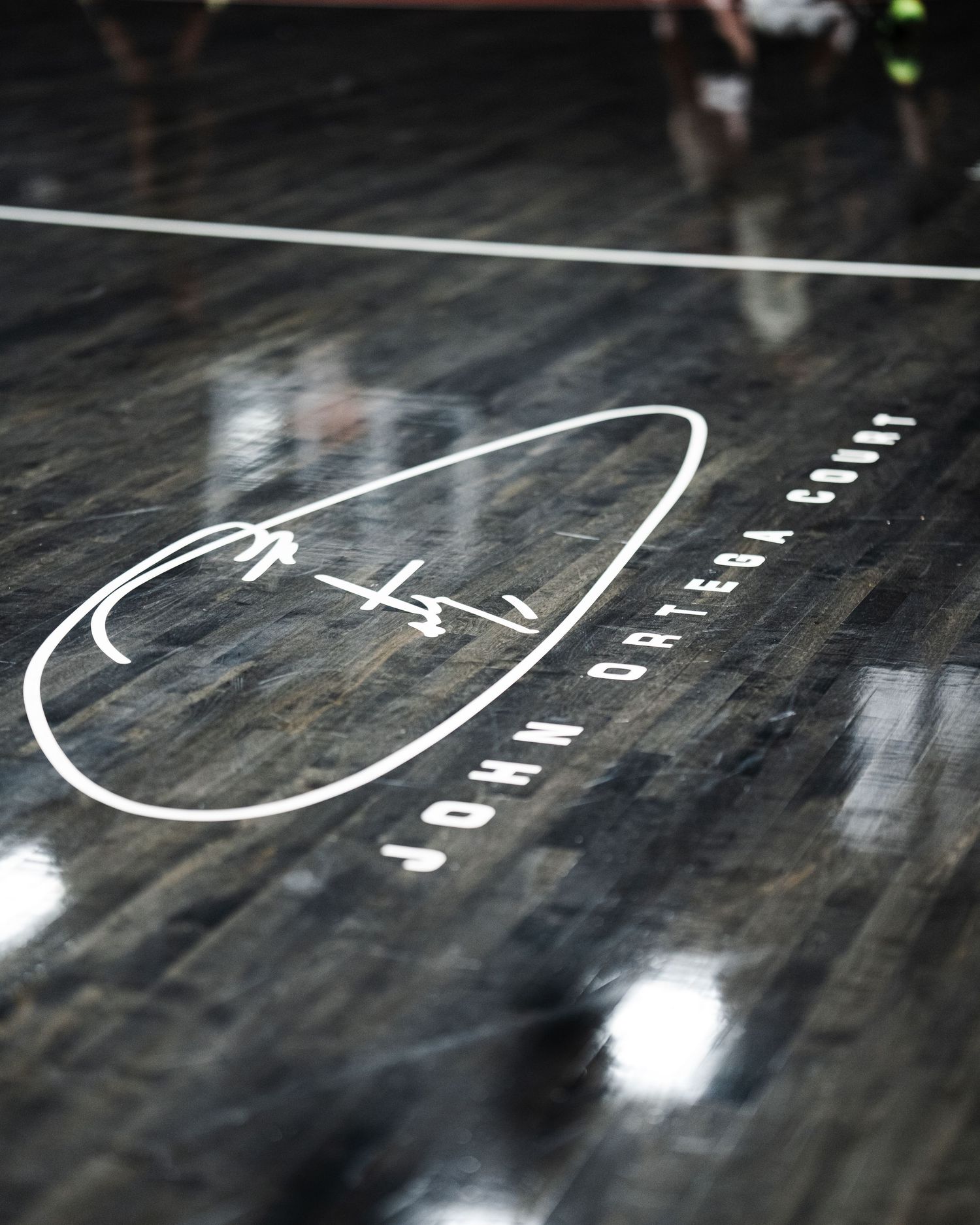 Close-up of the John Ortega Court logo engraved on a dark polished hardwood basketball floor.