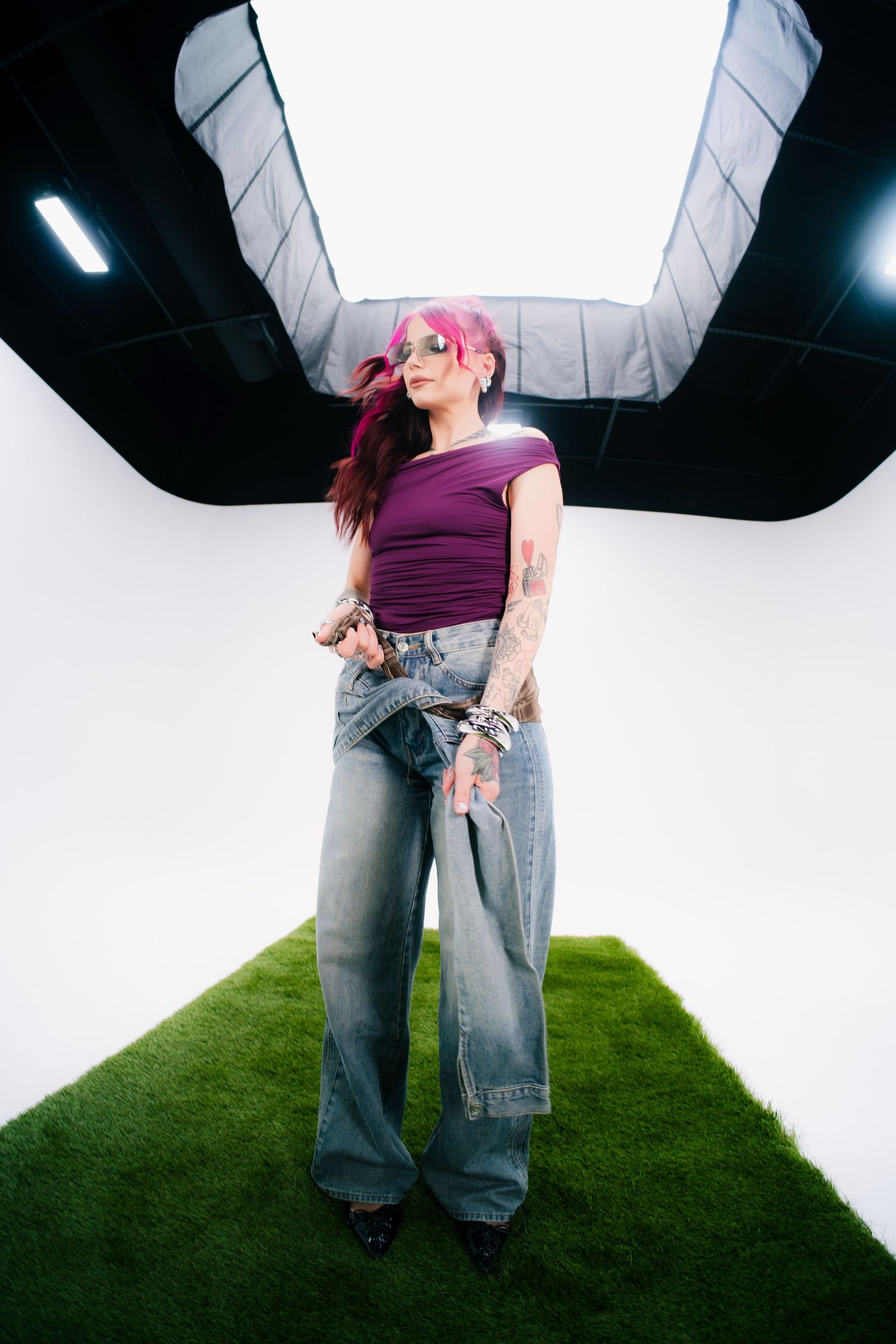 Woman with pink hair, tattoos, purple top, and wide-leg jeans posing in a white photo studio with green grass floor.