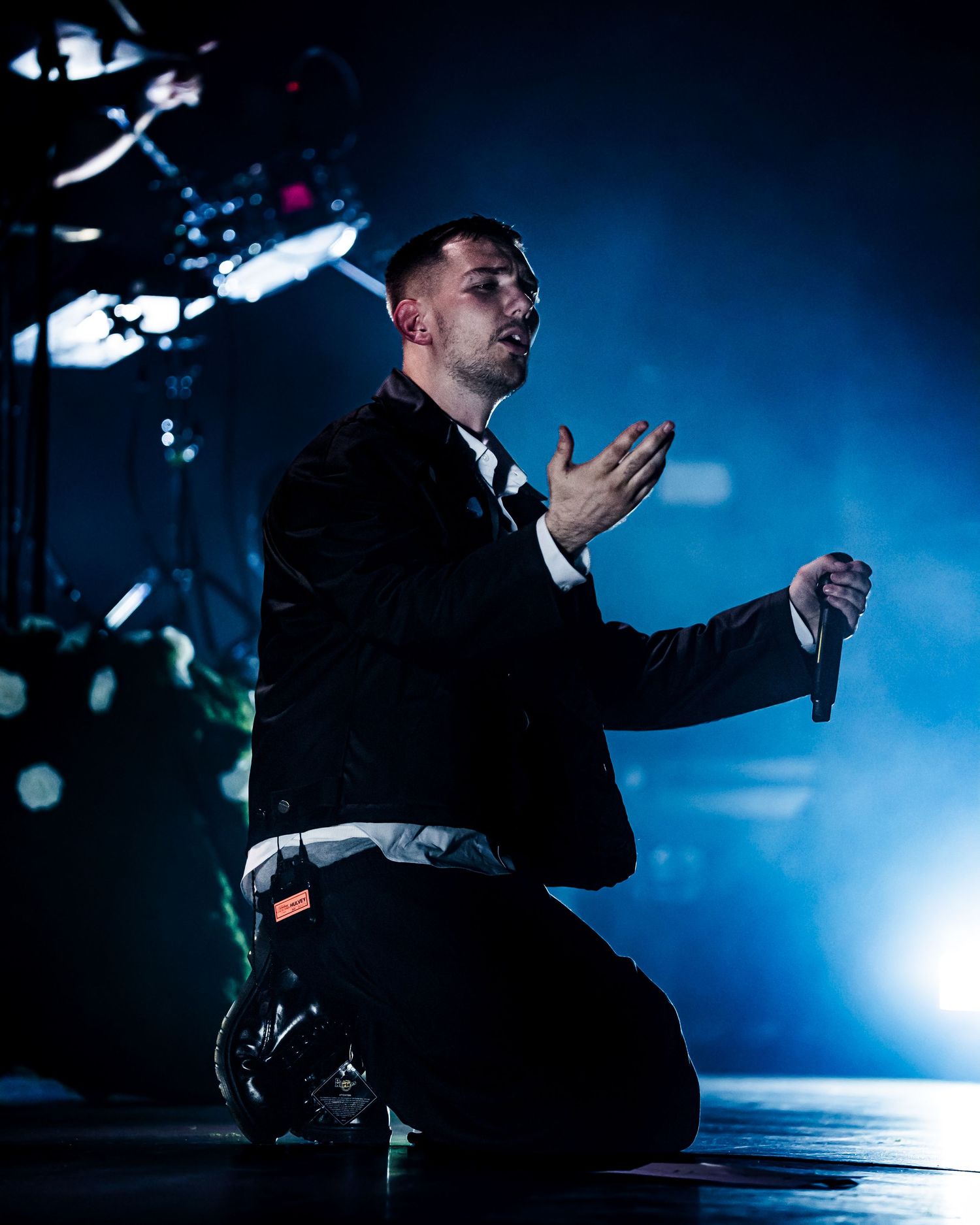 Male performer kneeling on stage in black suit, gesturing with hand under dramatic blue stage lighting.