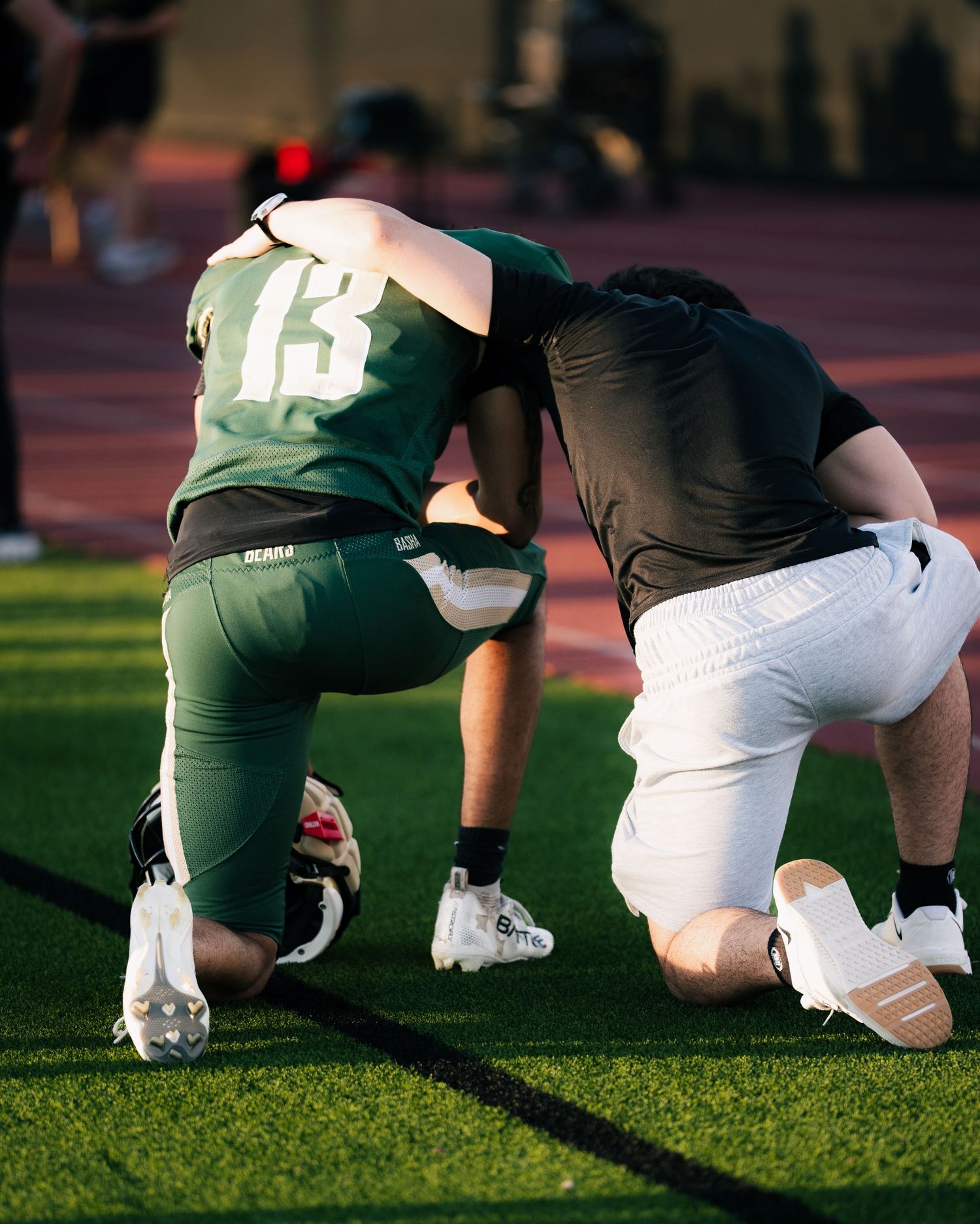 Two football players kneel together on a green turf field, one wearing jersey number 13 in green uniform.