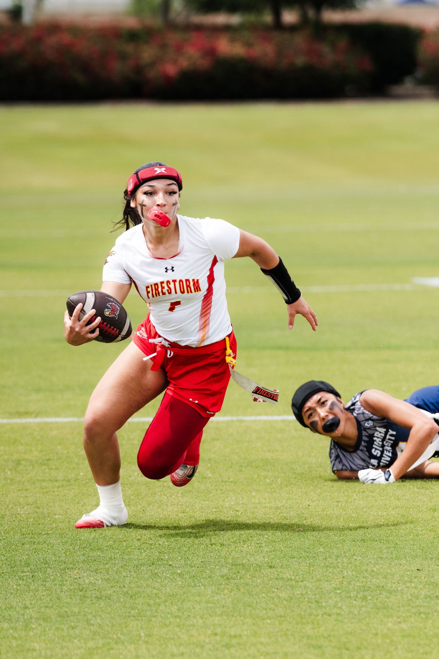 Female flag football player in Firestorm jersey runs with ball as defender falls behind her on green field.