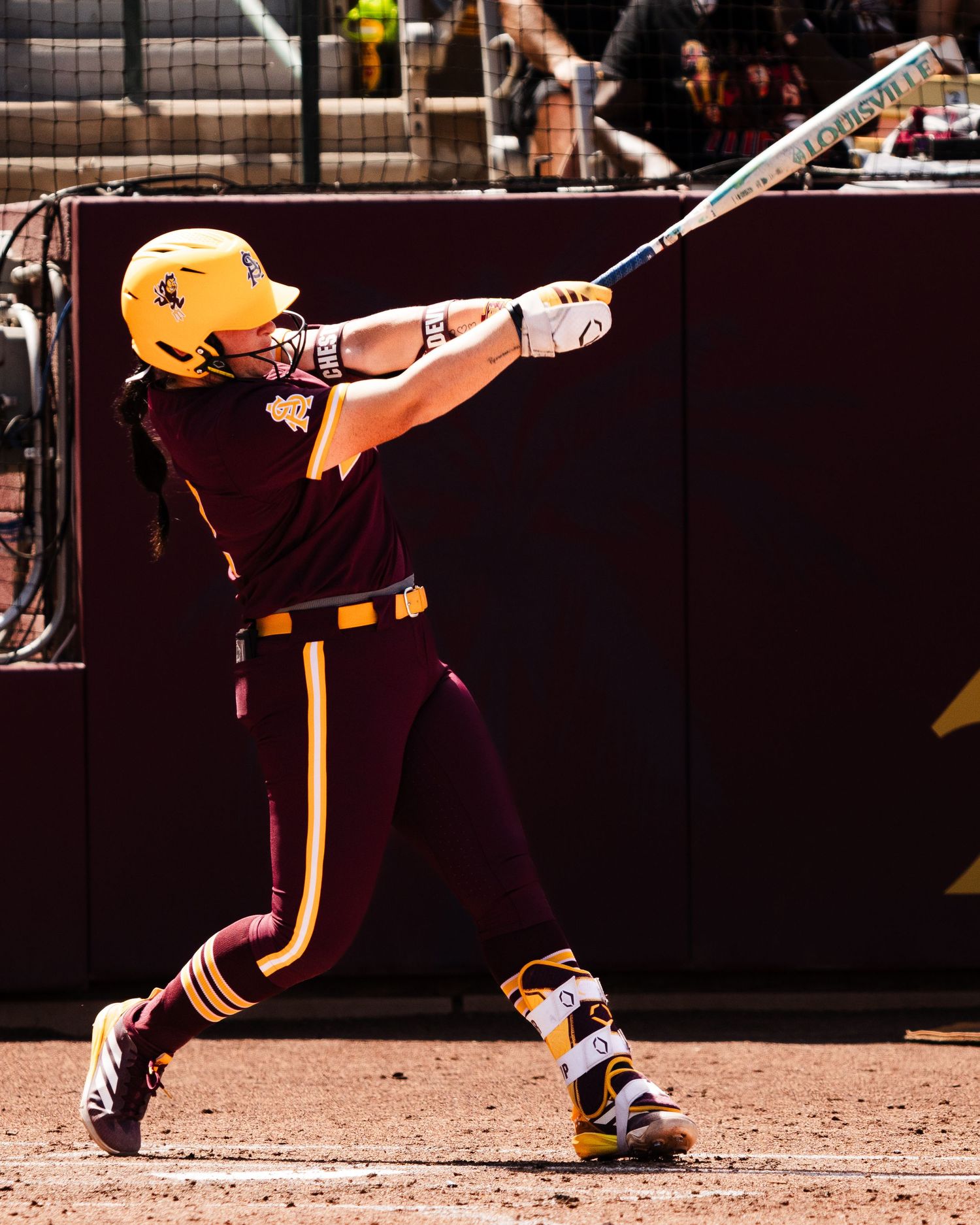 Arizona State Sun Devils softball player swings bat during a college game wearing maroon and gold uniform.