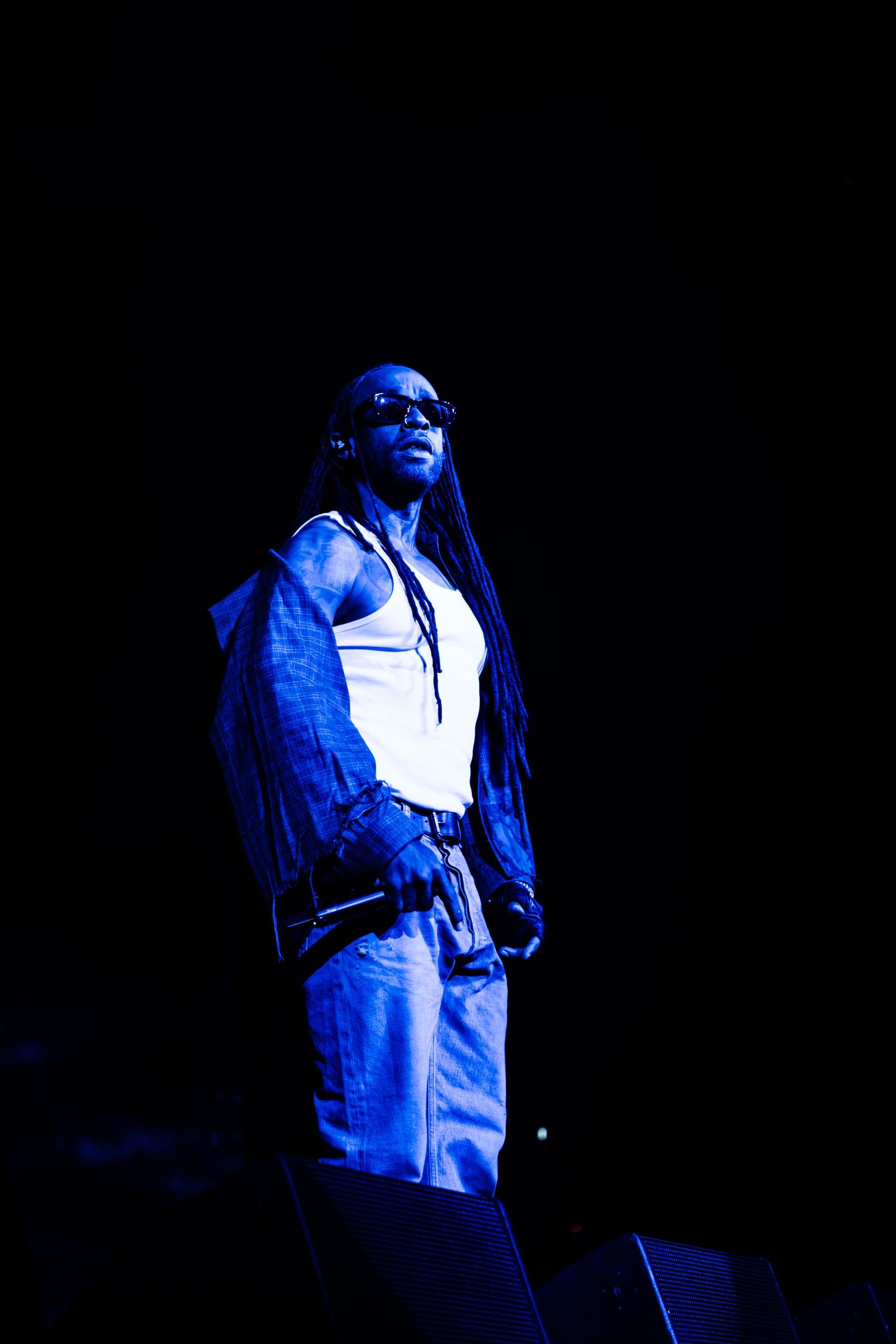 Performer in sunglasses, white tank top, and denim jacket with long locs holds a microphone on a dark stage under blue concert lighting.