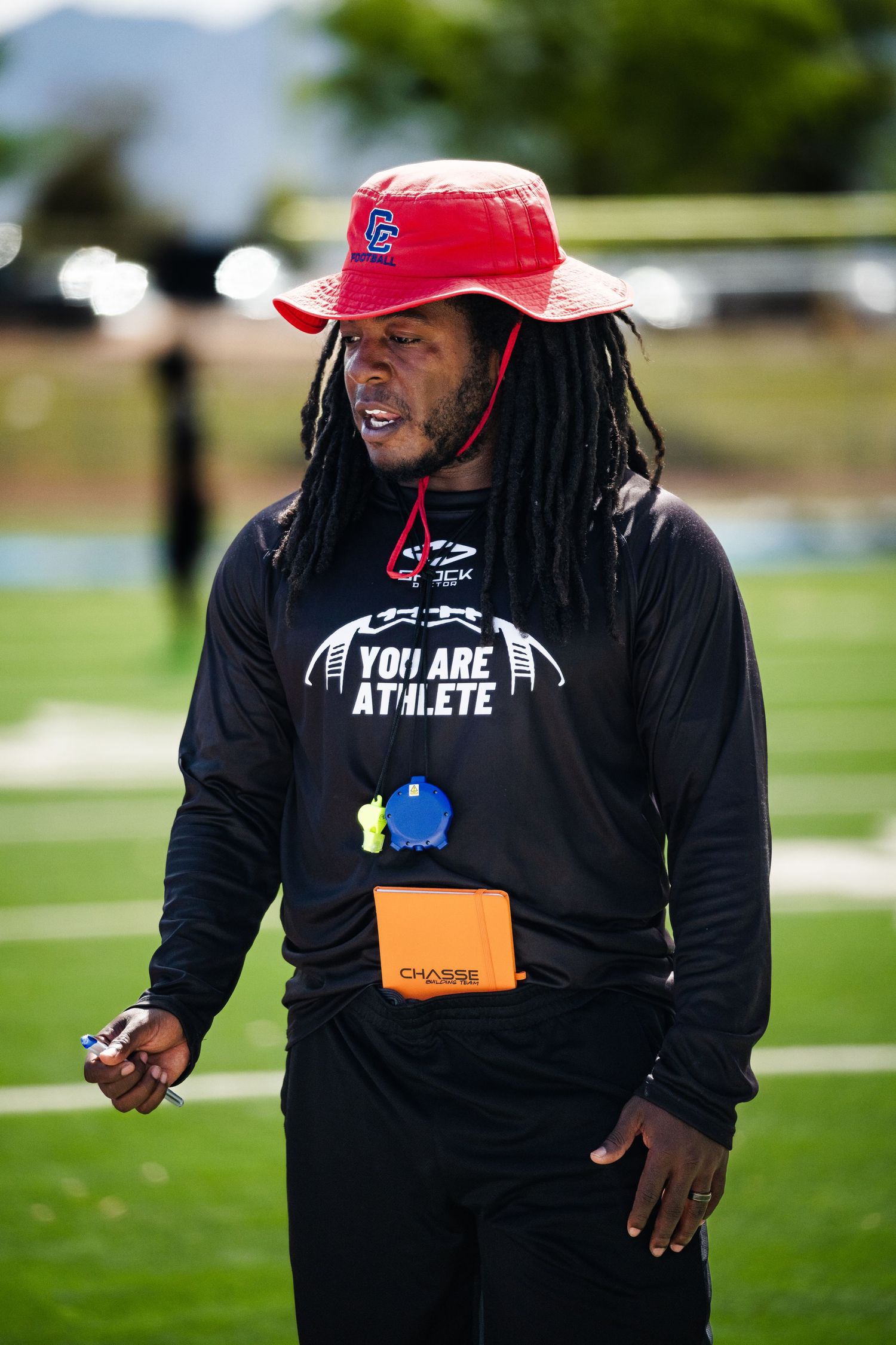 Coach wearing a You Are Athlete shirt and red bucket hat speaks during an outdoor football camp.
