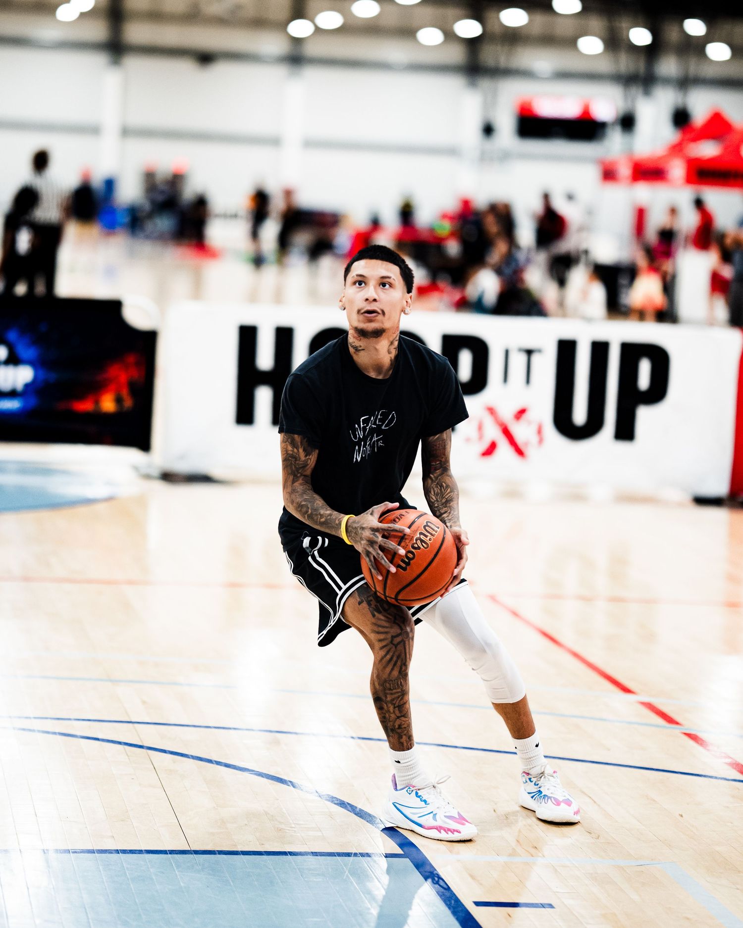 Tattooed basketball player in black jersey prepares to shoot at indoor Hoop It Up tournament event.