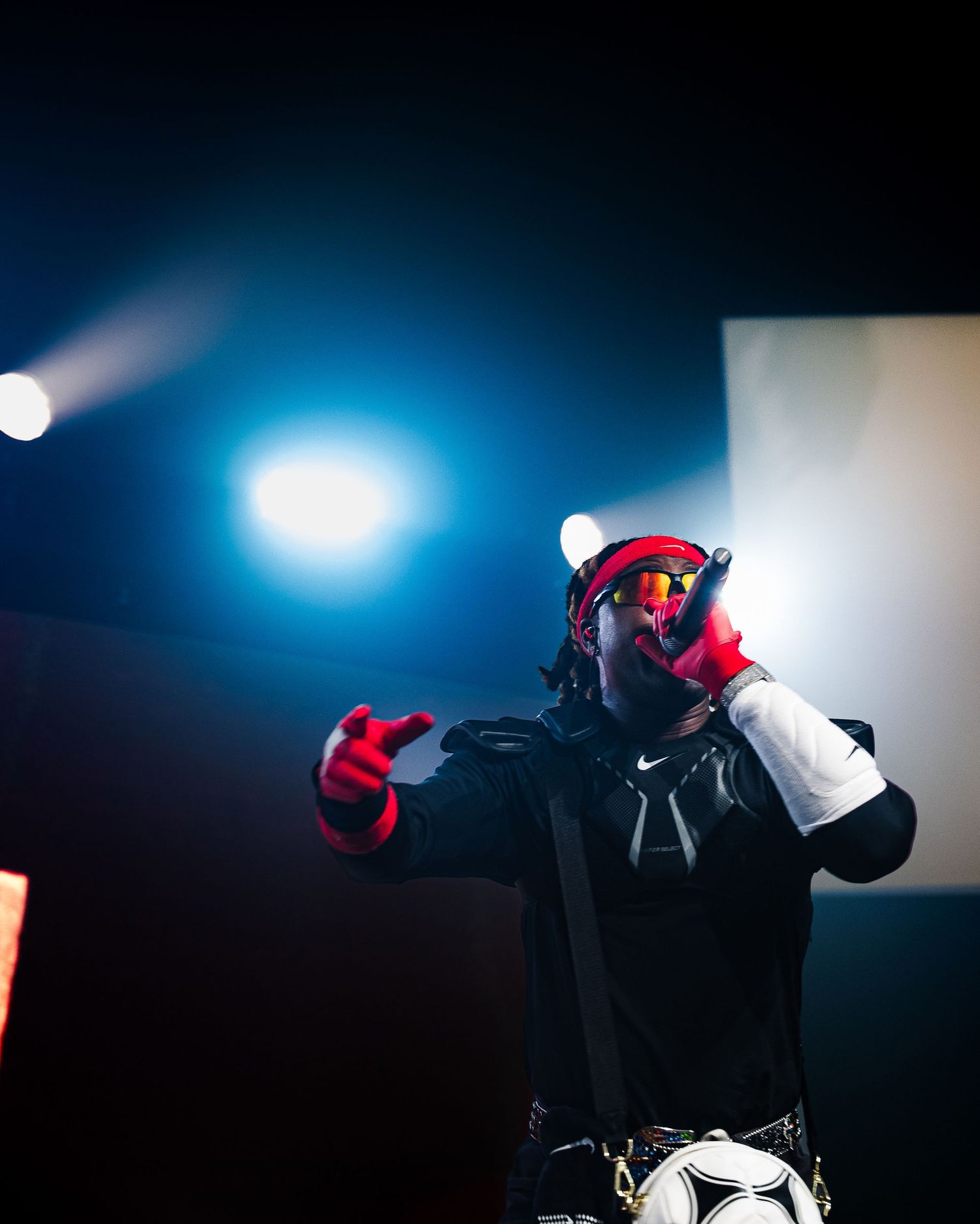 Rapper on stage wearing red goggles, red gloves, and black outfit performing under dramatic blue stage lighting.