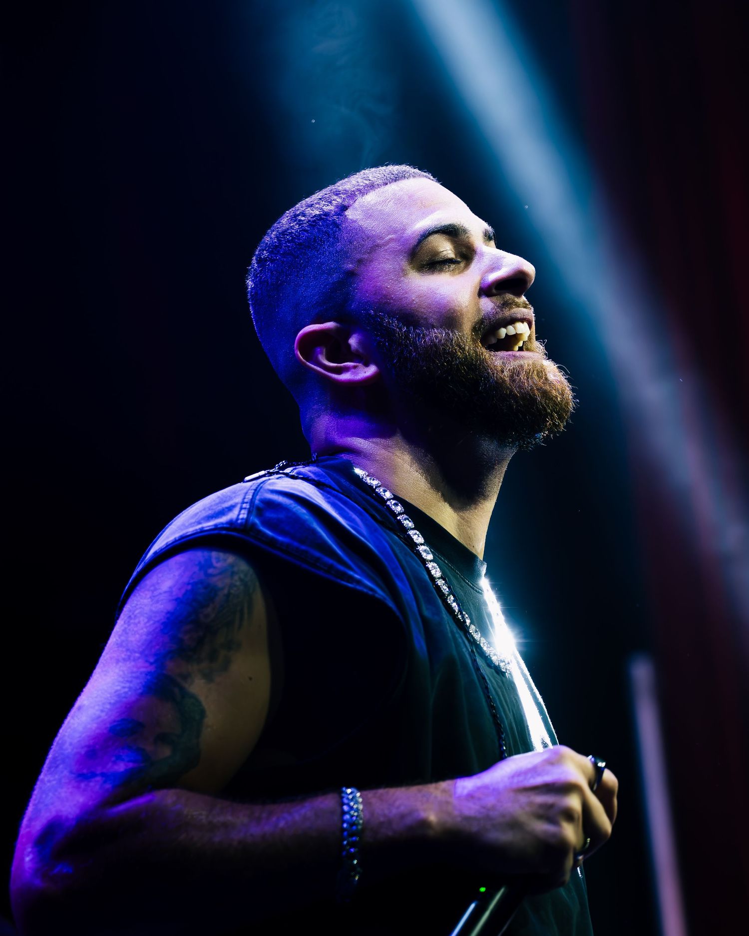 Smiling bearded man with tattoos performing on stage under purple and blue spotlights wearing a sleeveless black shirt.