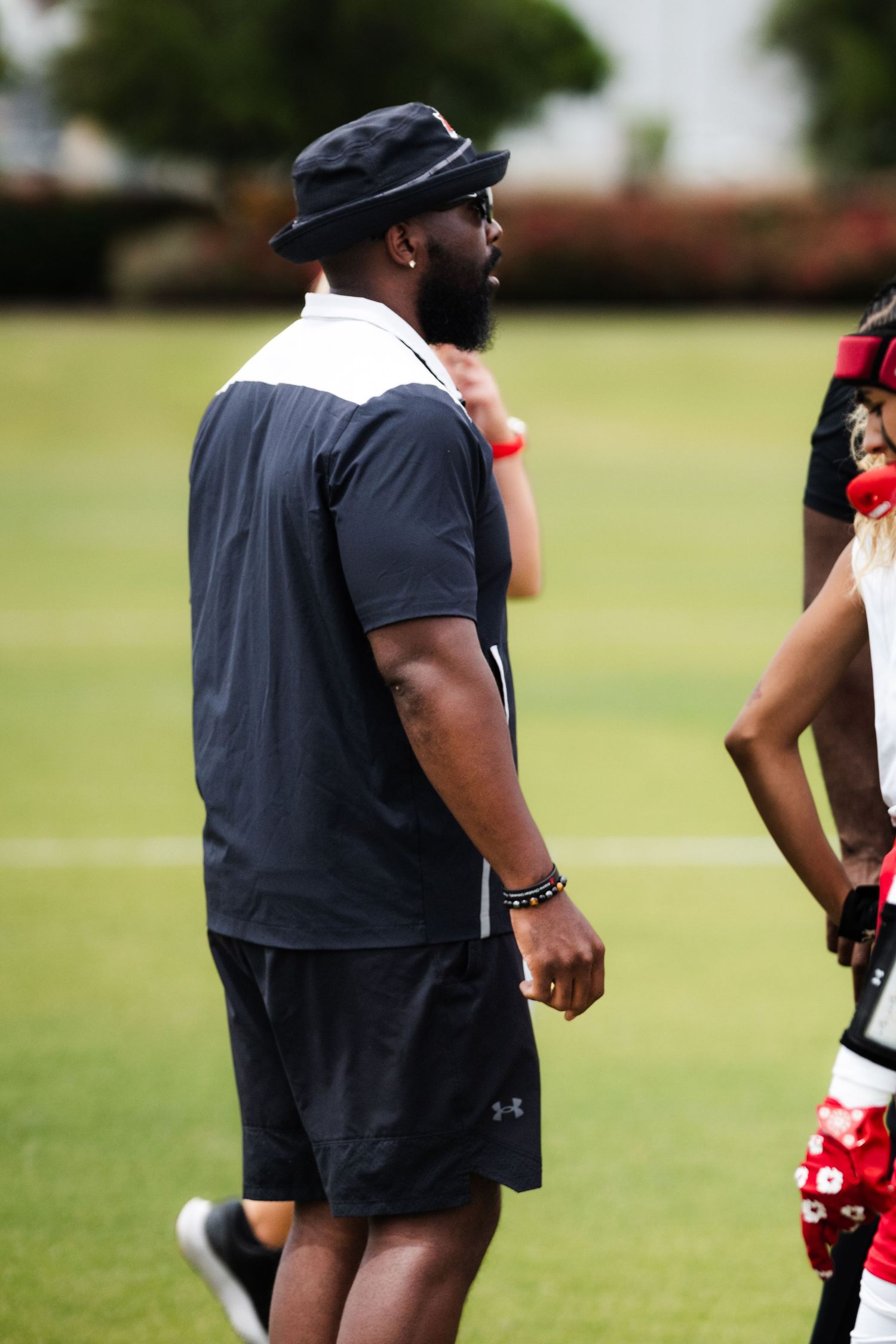 A bearded man in a black hat and shorts stands on a green sports field during an outdoor football practice session.