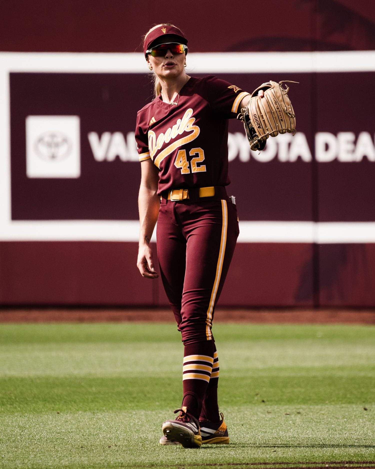 ASU softball player #42 in maroon uniform raises glove near outfield wall.