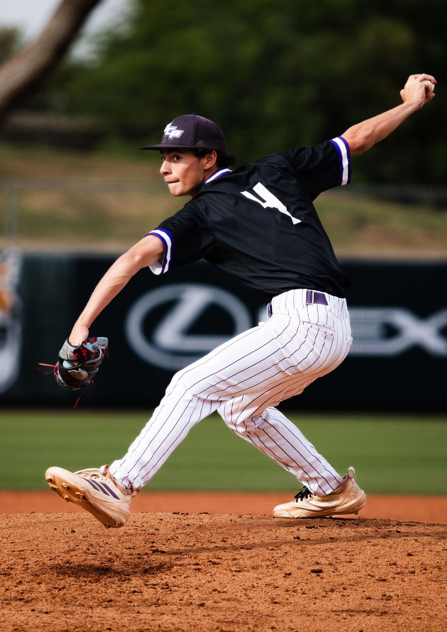 Baseball pitcher in black pinstripe uniform mid-windup on dirt mound during game with Lexus advertisement in background.