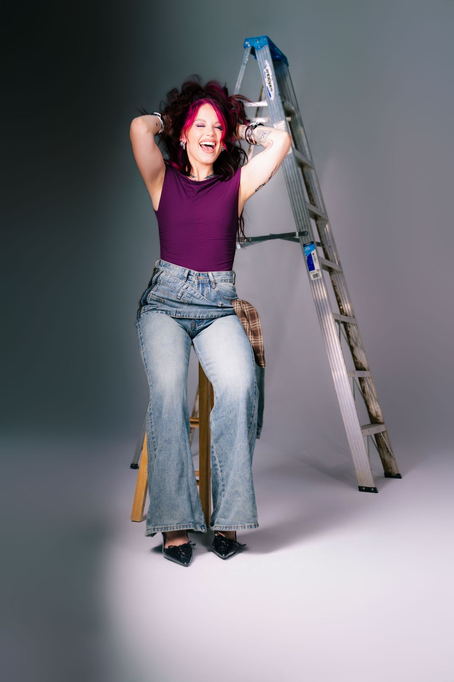 Woman with pink hair laughing in purple top and wide-leg jeans, posing beside a metal ladder on gray background.