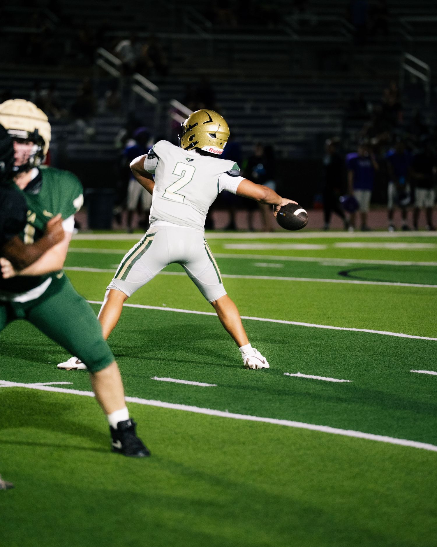 Football player number 2 in white uniform throws a pass during a night game on a green field.