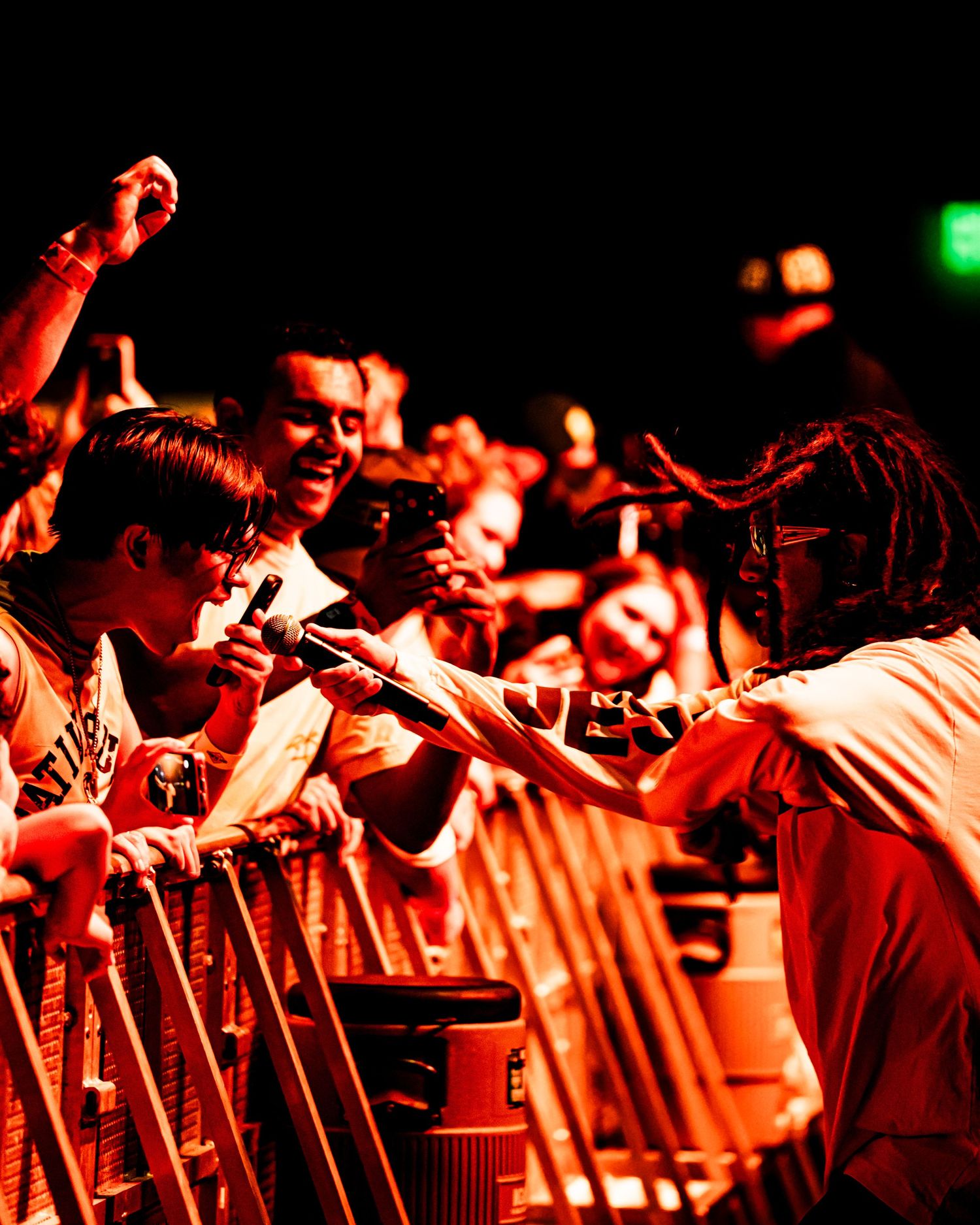 Artist with dreadlocks reaches out to excited fans over barrier under red concert lighting.