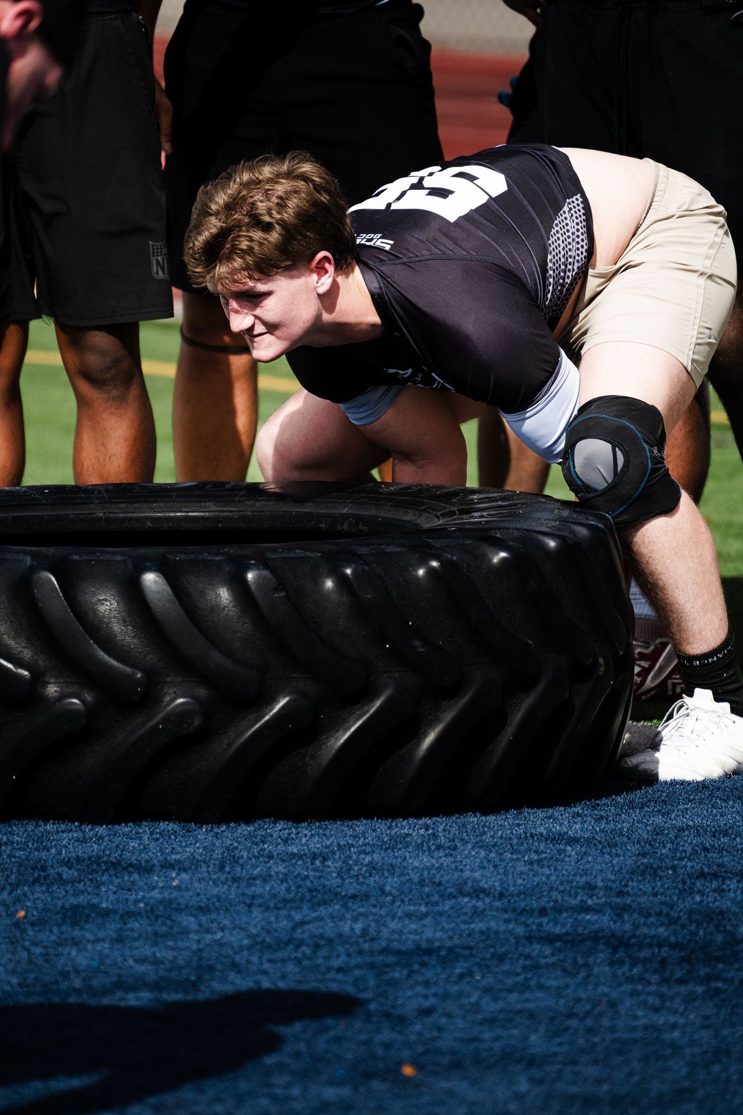 Young athlete flips large black training tire on blue turf during football strength conditioning drill.
