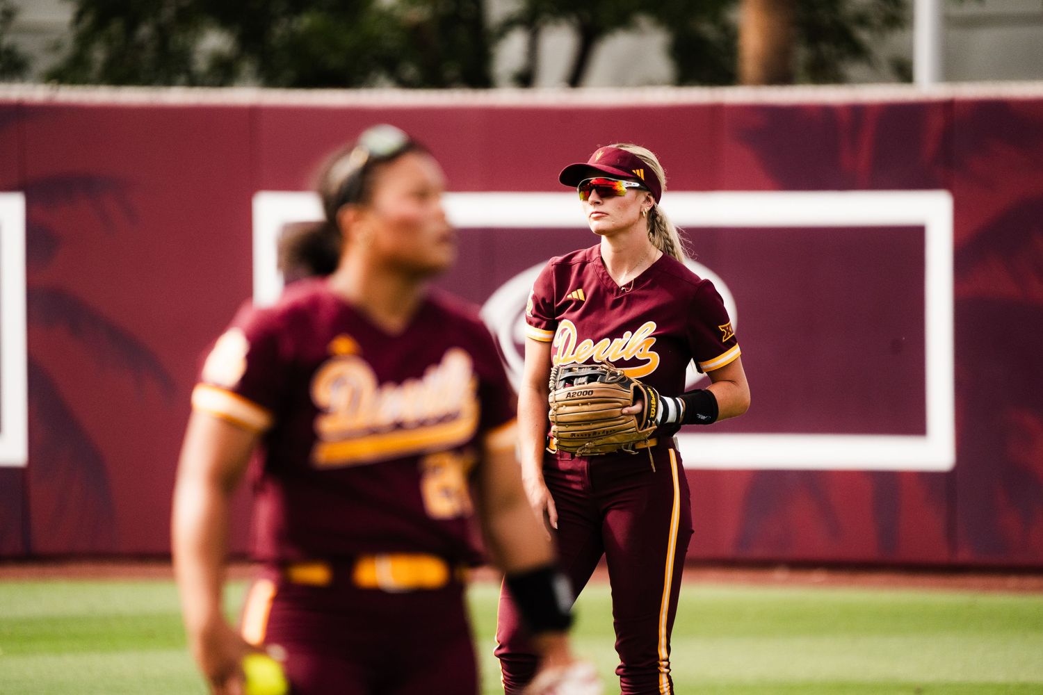 Two Arizona State Sun Devils softball players on the field during a game, wearing maroon and gold uniforms.