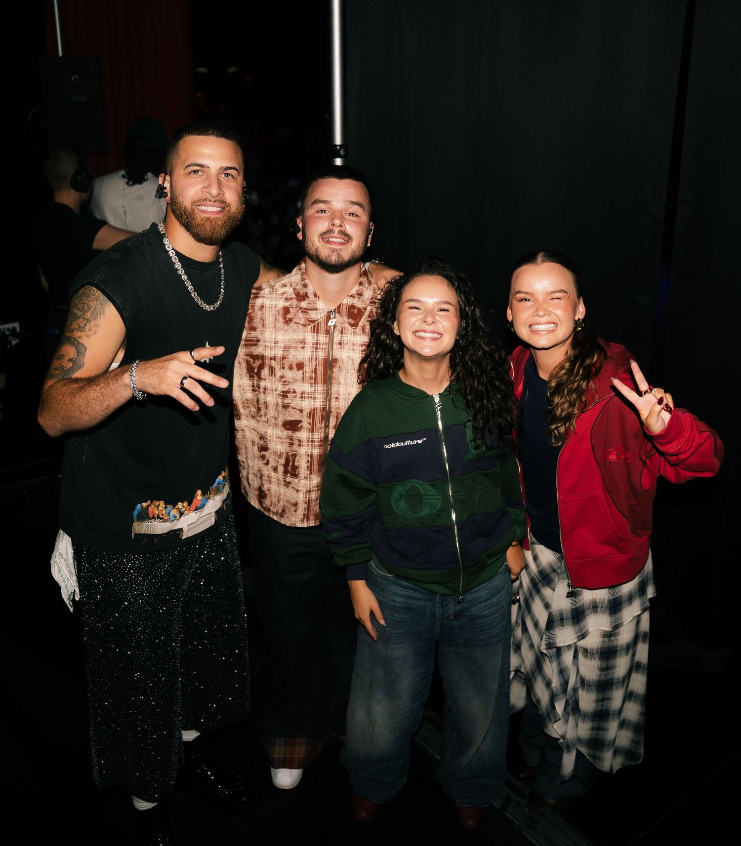 Four friends pose together smiling in a dark venue, wearing casual colorful outfits, one flashing a peace sign.
