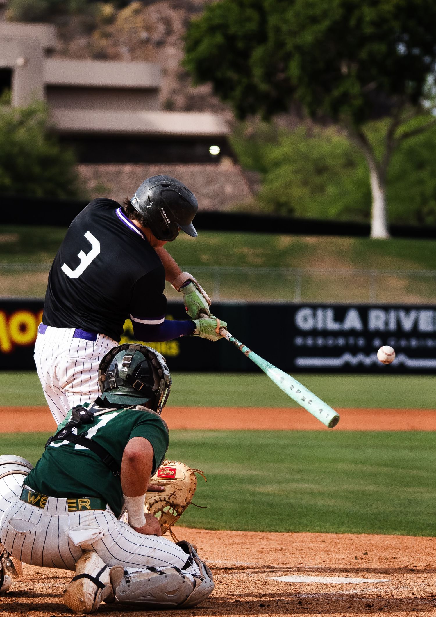 Baseball player number 3 makes contact with the ball as the catcher crouches at Gila River stadium.