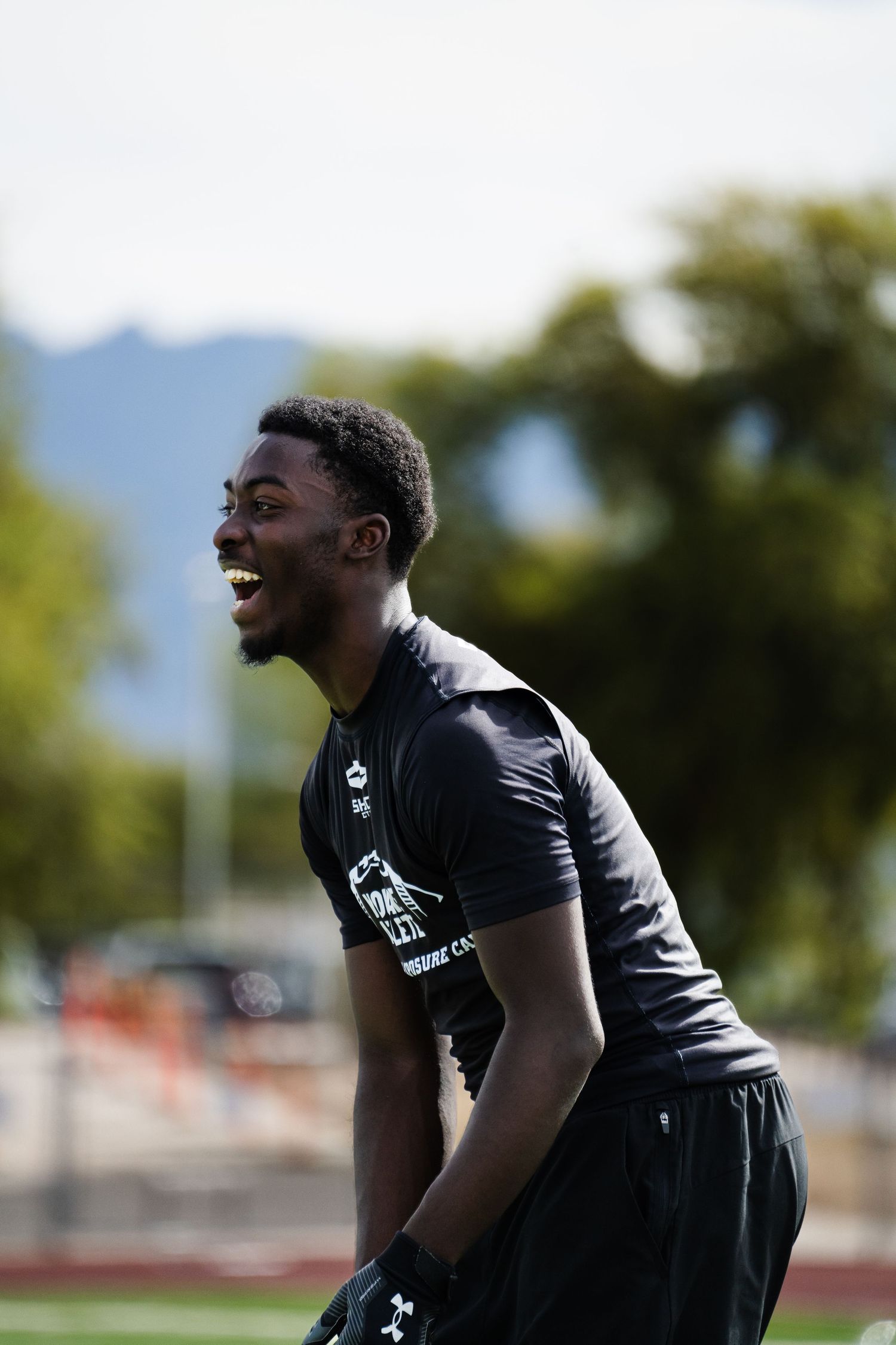 Smiling young athlete in black Shock Doctor jersey laughing during outdoor football combine on sunny day.