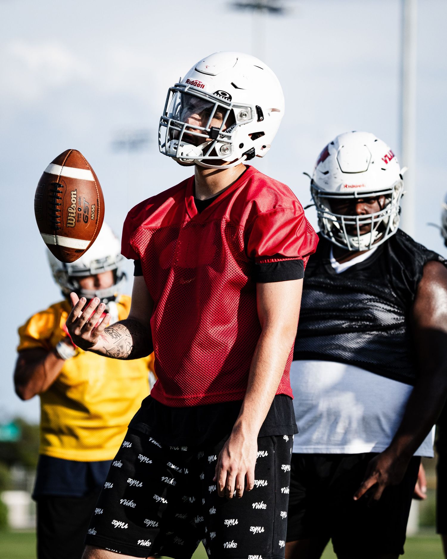 Football player in red jersey and helmet catches a Wilson football during an outdoor practice session with teammates.