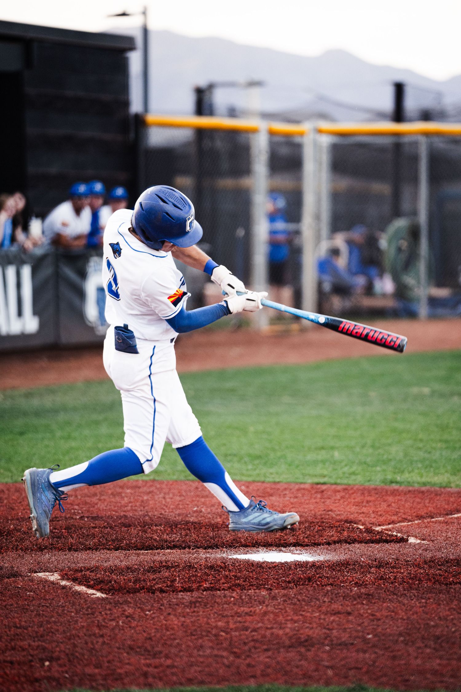 Baseball batter in white and blue uniform swings at pitch during a game on a red dirt infield.