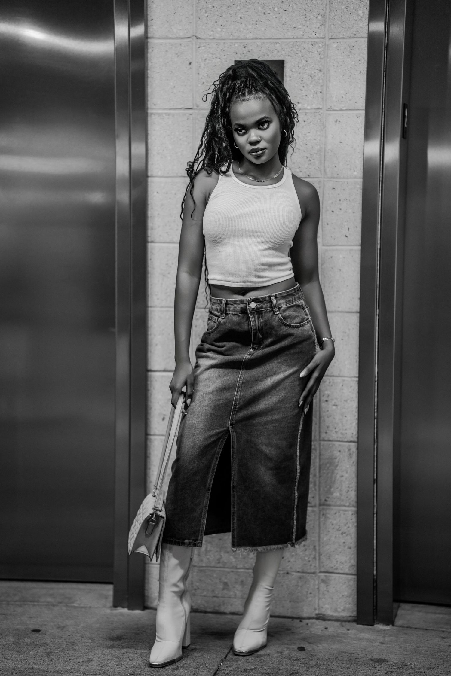 Black and white photo of a young woman in a white tank top, denim midi skirt, and white boots posing near elevators.