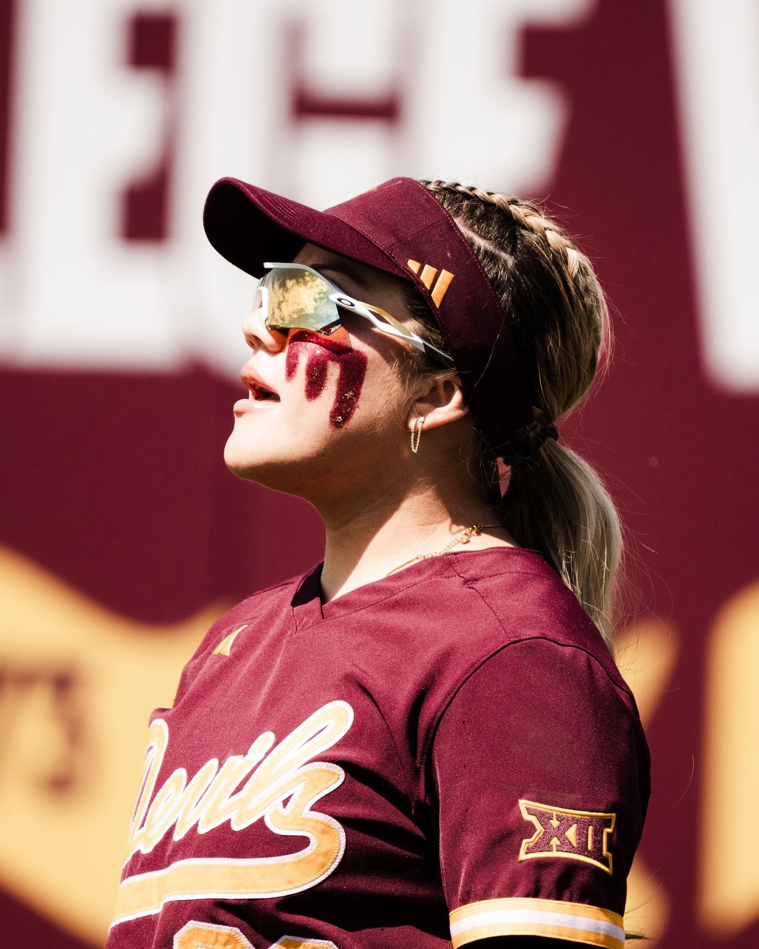 Female softball player in maroon Big 12 uniform with face paint, wearing gold sunglasses and cap, looking upward outdoors.