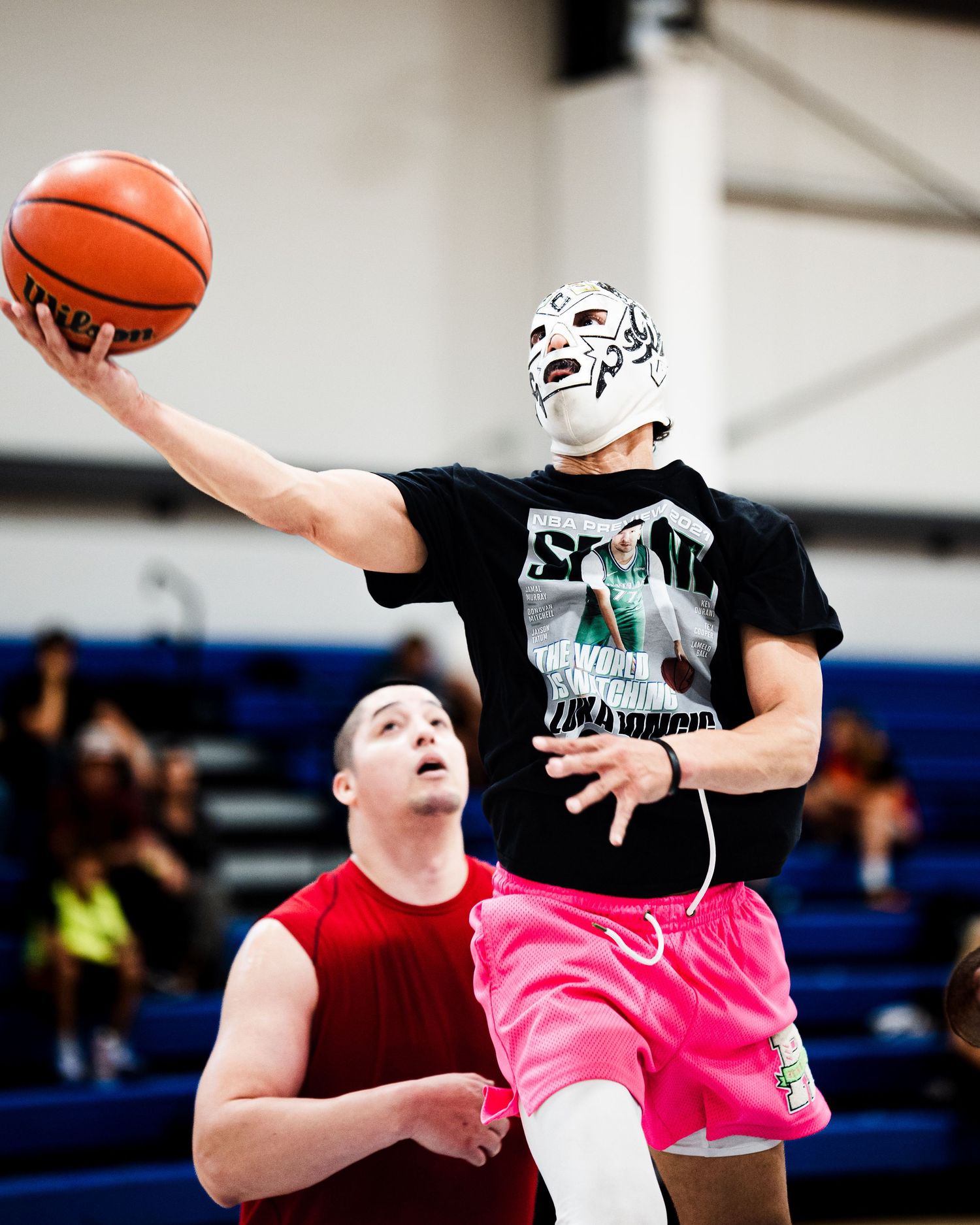 Player wearing a wrestling mask leaps for a layup during an indoor basketball game while defender watches.