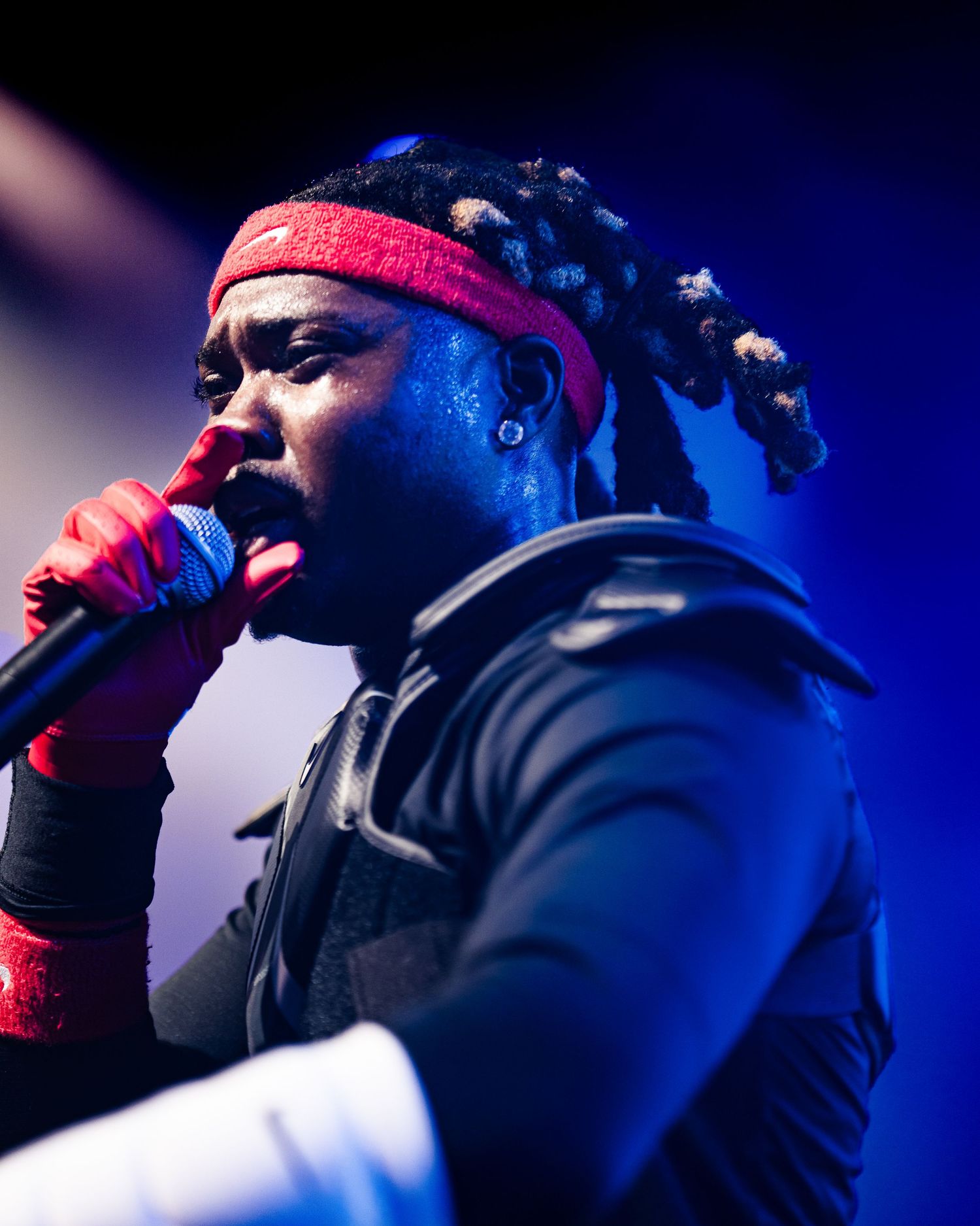 A male rapper with dreadlocks and red headband performs on stage under dramatic blue stage lighting.