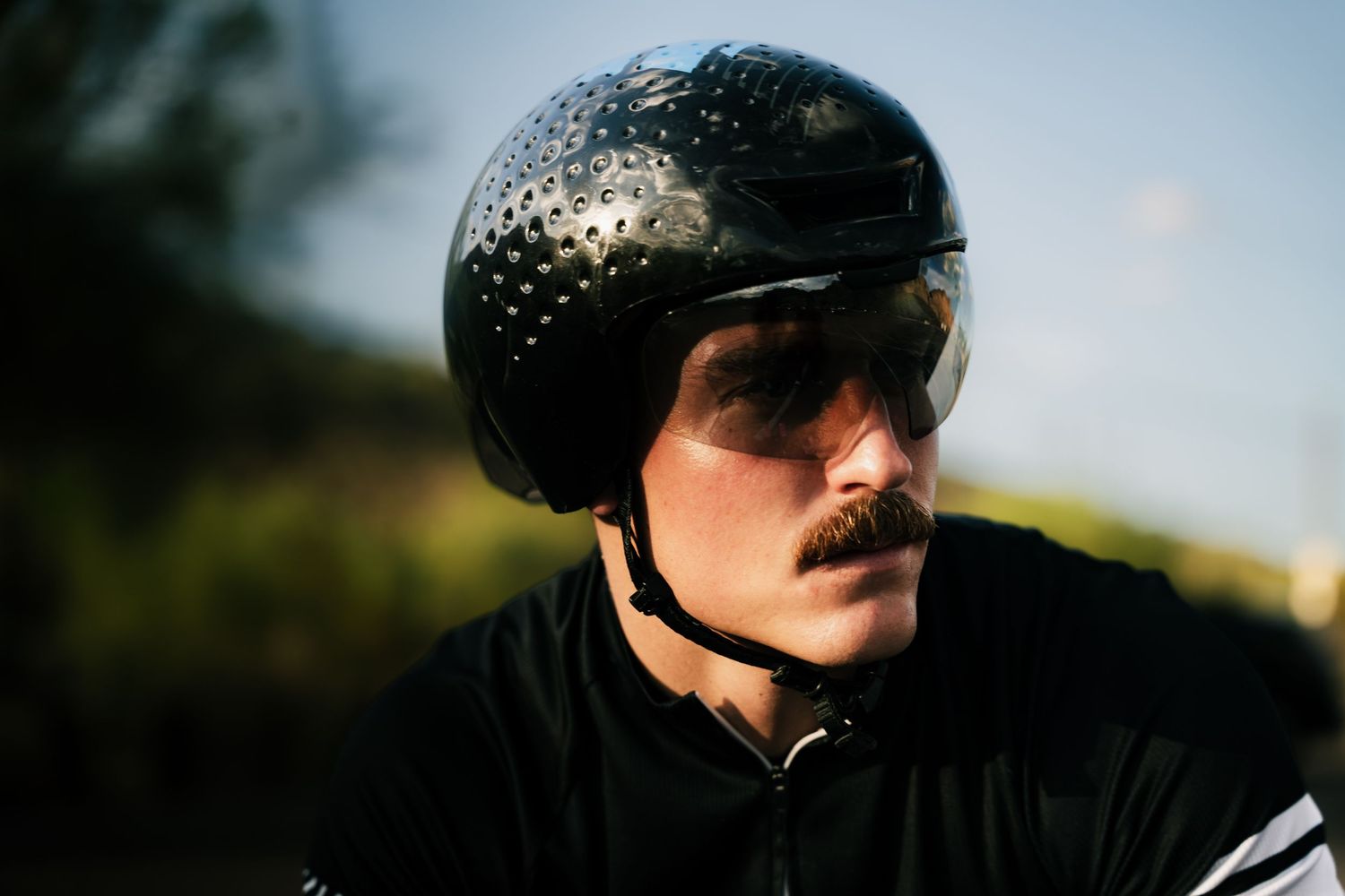Close-up of a male cyclist wearing a black perforated helmet and sunglasses outdoors.